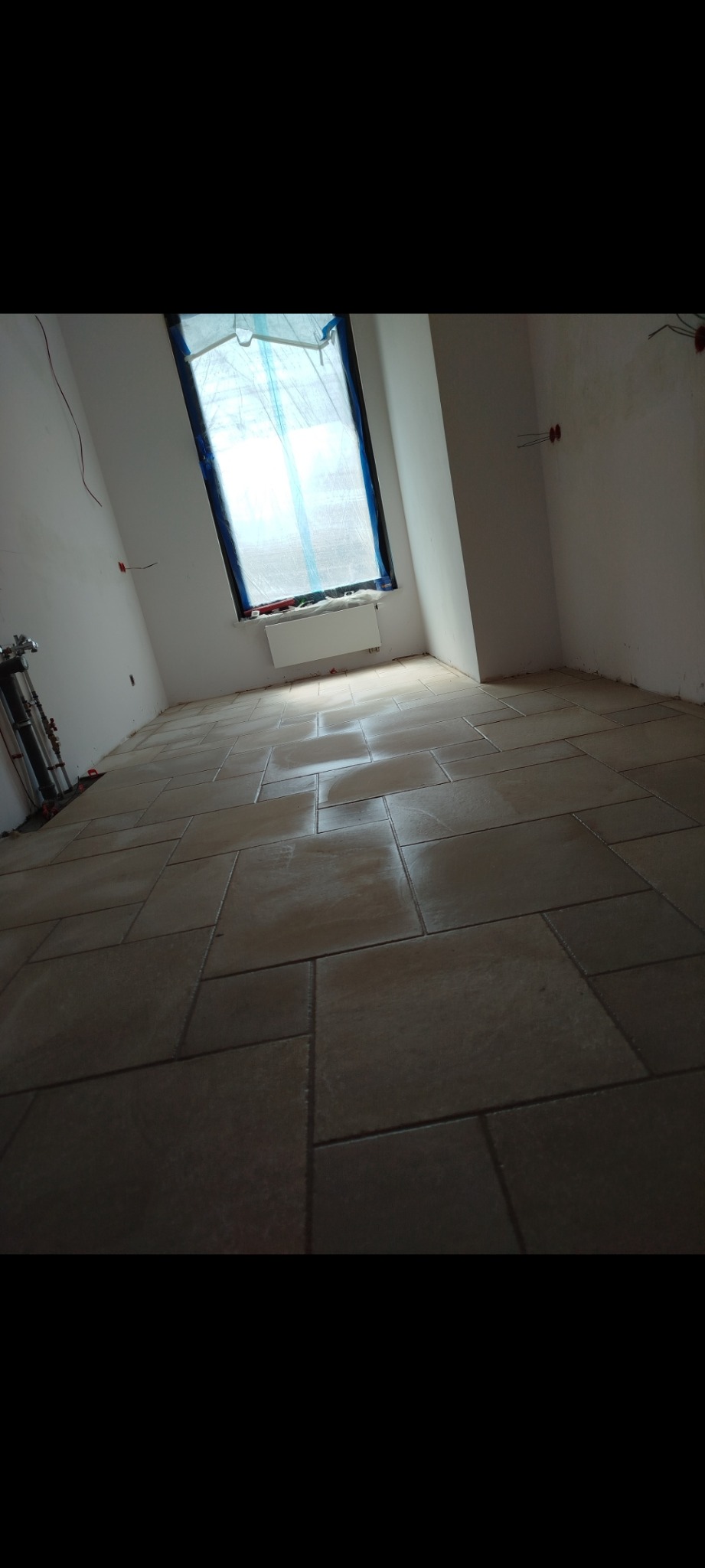 Ułożone płytki podłogowe w jasnym odcieniu beżu w pokoju z oknem zabezpieczonym folią malarską. Widoczne przewody elektryczne i rury instalacyjne.