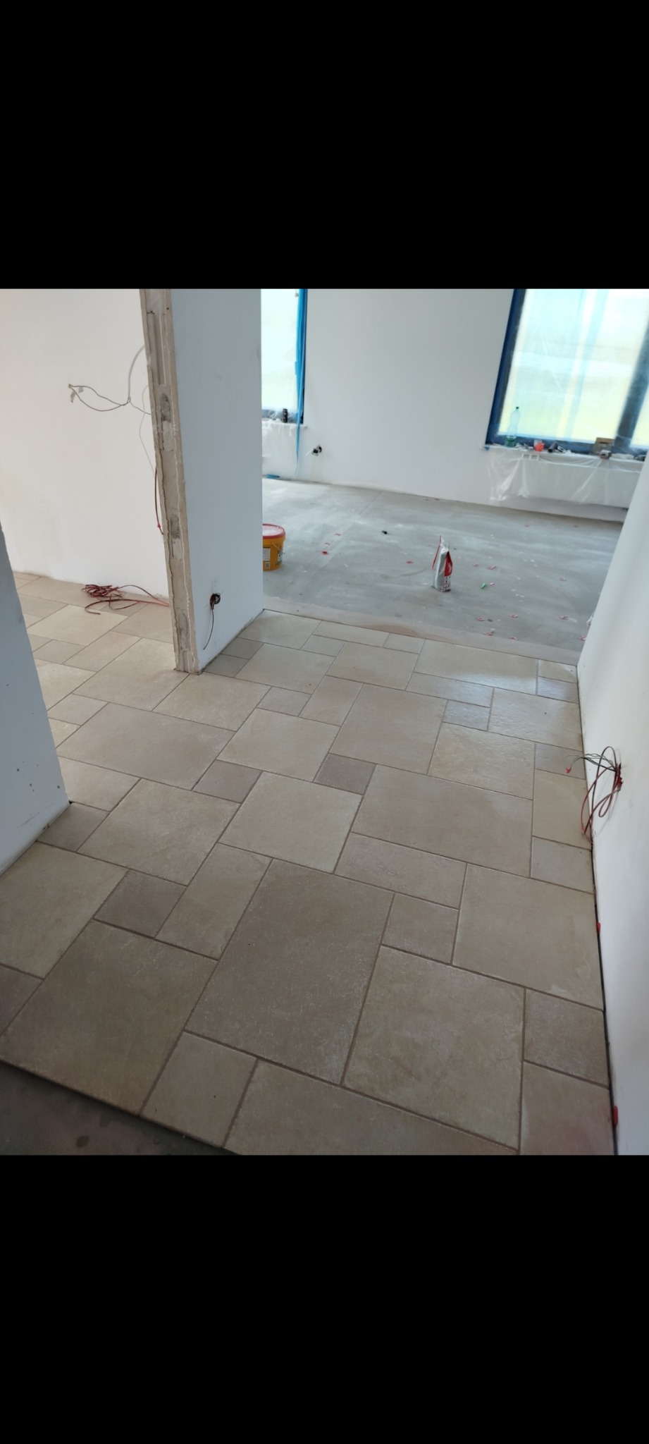 Ukończona podłoga z beżowych płytek ułożonych w geometryczny wzór. Widoczne białe ściany w trakcie wykończenia i okno z niebieską folią ochronną.