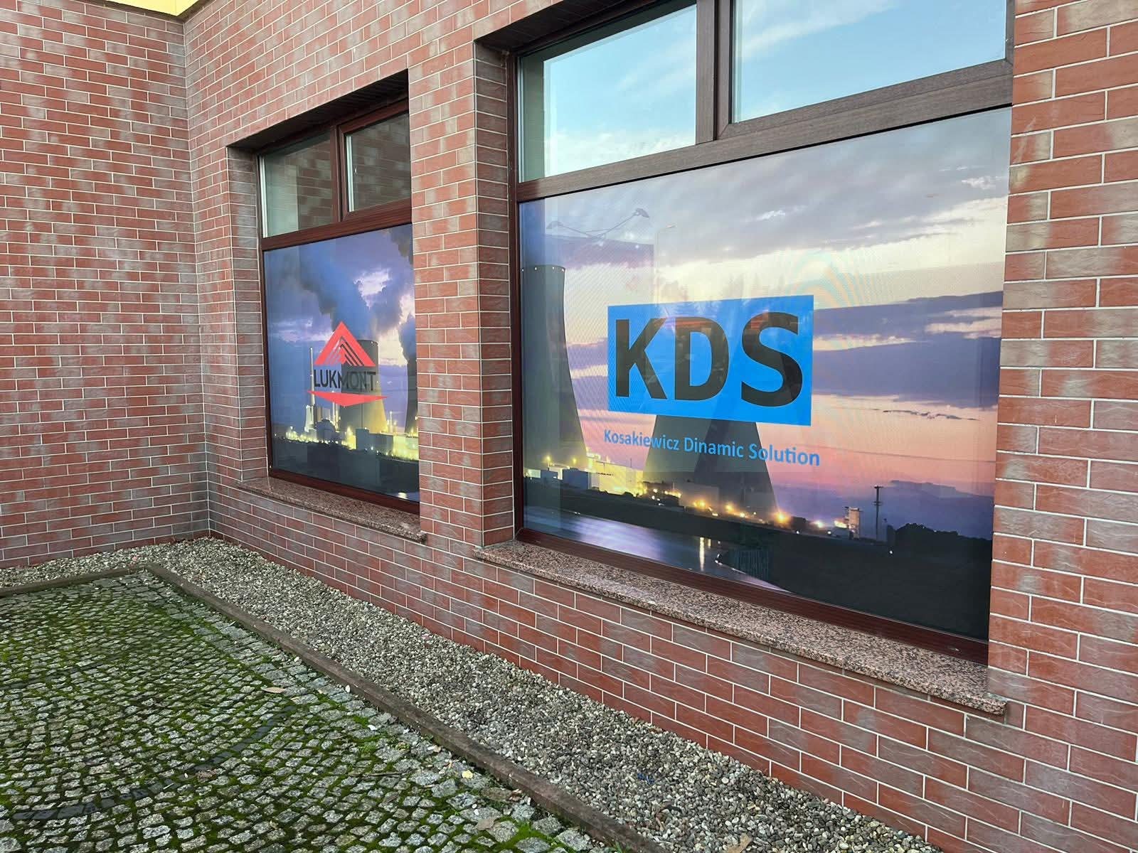 Oklejone folią okna budynku z cegły. Na szybach widoczne logotypy firm 'Lukmont' i 'KDS' z grafiką przemysłowego krajobrazu w tle, w brązowych ramach.
