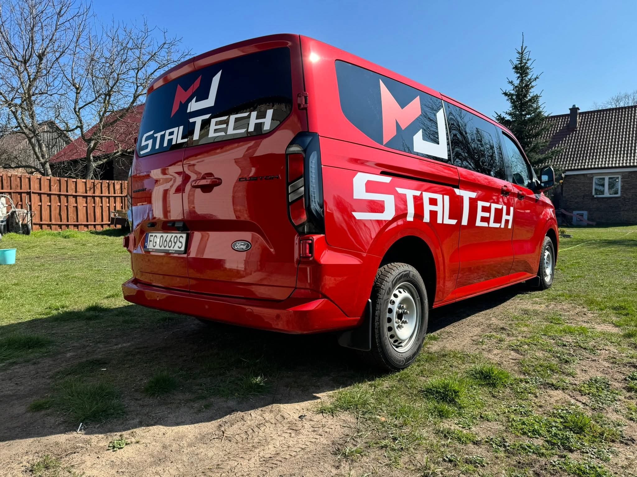 Czerwony Ford Transit z logo 'MJ STALTECH' na oknach i karoserii, zaparkowany na trawie w słoneczny dzień. Widoczny numer rejestracyjny i elementy nadwozia.