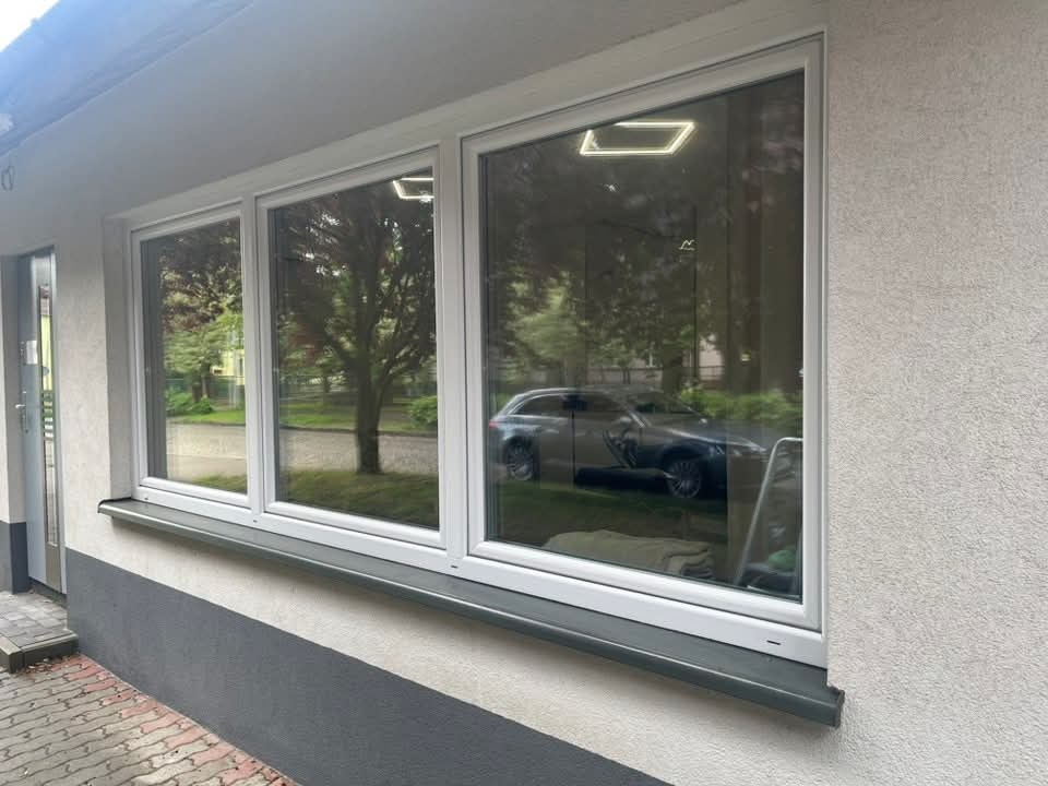 Okno z folią przeciwsłoneczną, odbijające drzewo i samochód, zamontowane w budynku z elewacją w odcieniach szarości i bieli. Widok z zewnątrz, w dzień.