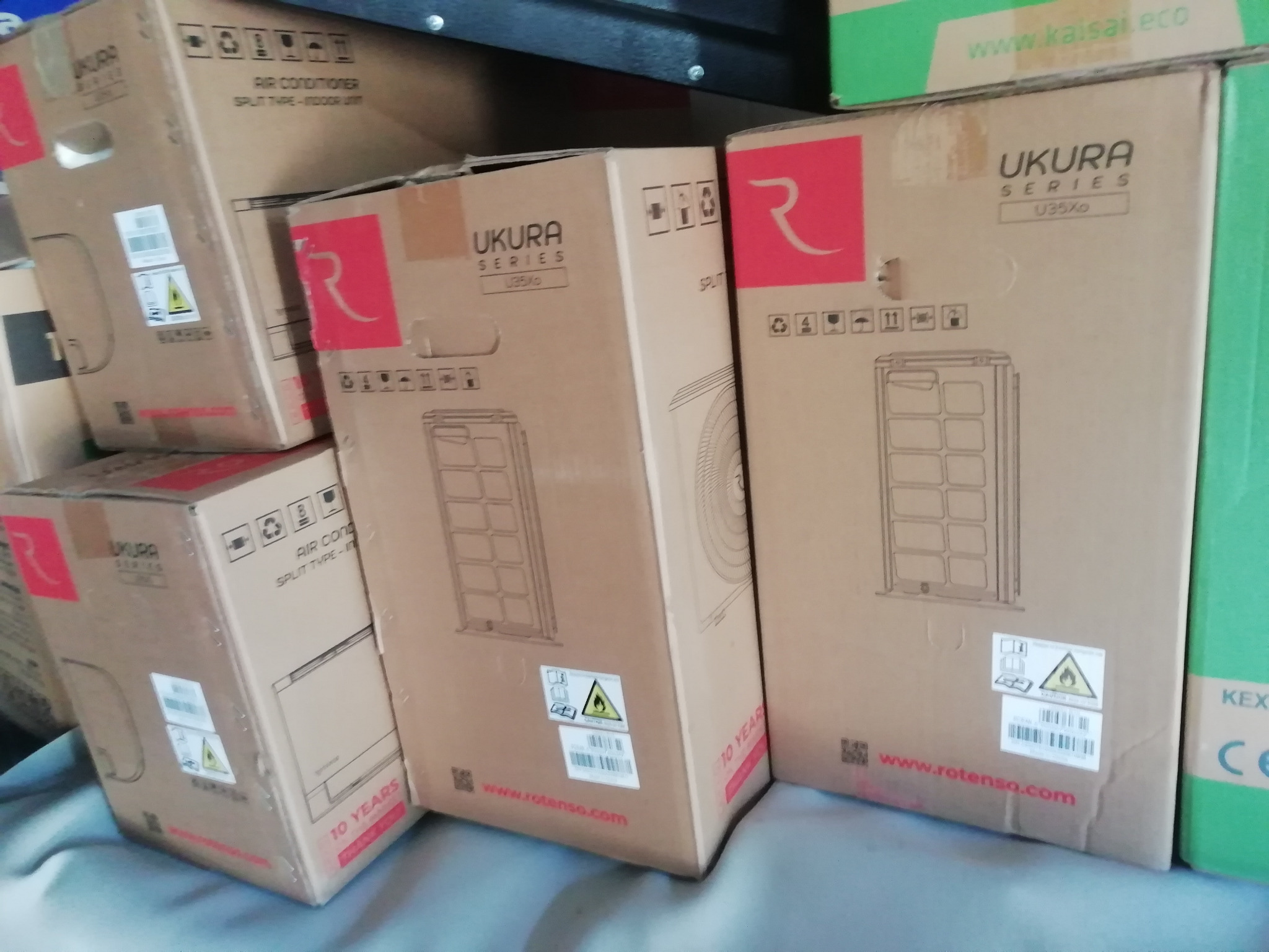 Stos kartonów z klimatyzatorami Ukura Series U35Xo, z widocznymi grafikami i oznaczeniami. Jeden karton częściowo zasłonięty zielonym opakowaniem Kaisai Eco.