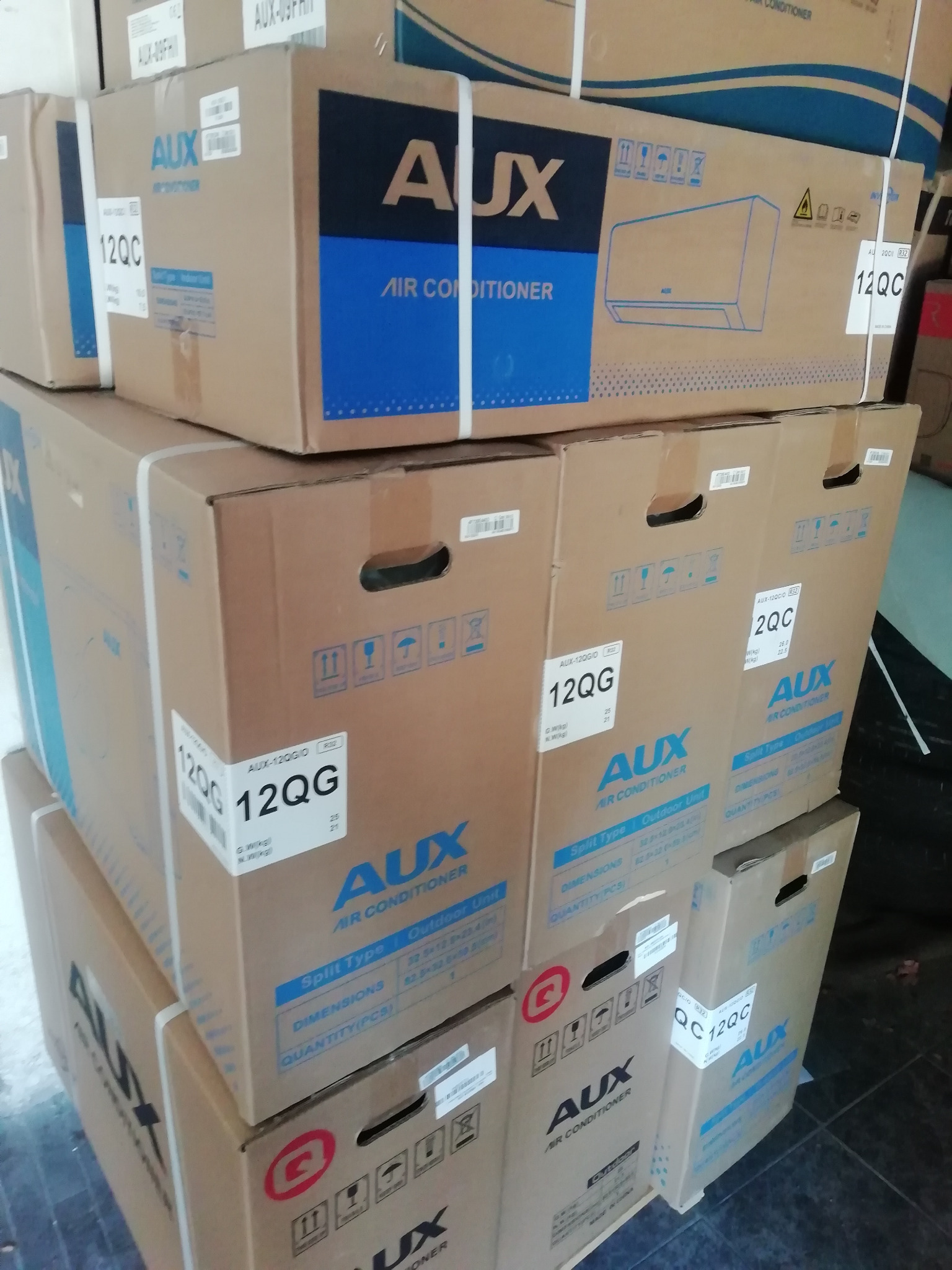 Stos kartonów z klimatyzatorami AUX, modele 12QC i 12QG, z widocznymi oznaczeniami i specyfikacjami technicznymi na opakowaniach. Widok z bliska, magazynowane produkty.