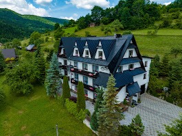 Pensjonat w górskiej scenerii. Trzy kondygnacje, ciemny dach z lukarnami, białe ściany, balkony z kwiatami. Zielone wzgórza w tle, malowniczy krajobraz.