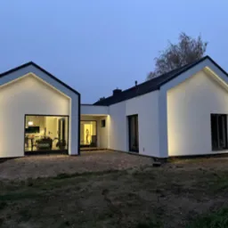 Nowoczesny, parterowy dom z białymi elewacjami i czarnym dachem, oświetlony subtelnym, ledowym światłem, widoczny o zmierzchu.