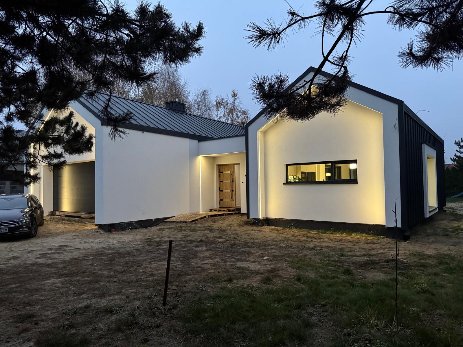 Nowoczesny dom jednorodzinny z białą elewacją i czarnym dachem, oświetlony delikatnym światłem, z widocznym garażem i fragmentem samochodu.