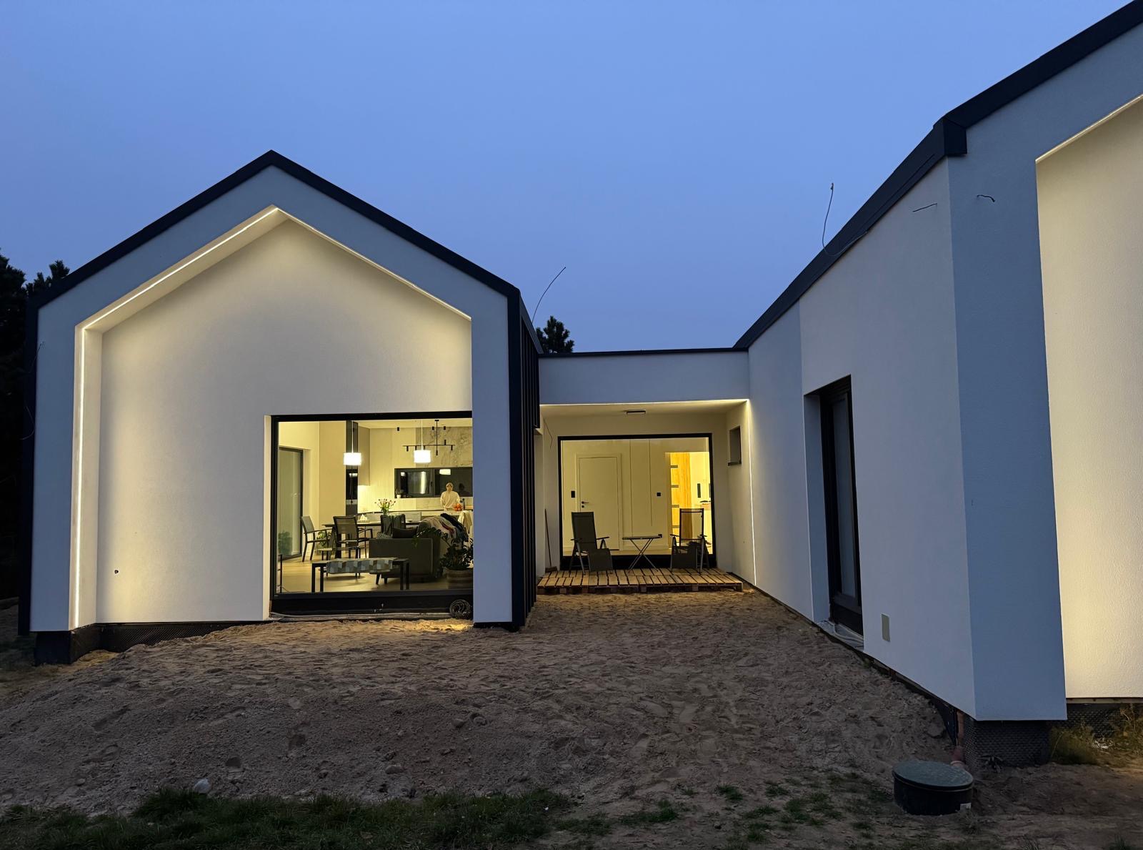 Nowoczesny dom z białymi ścianami i czarnym dachem, oświetlony od zewnątrz, widok na wnętrze przez duże okna. Teren wokół domu jeszcze niezagospodarowany.