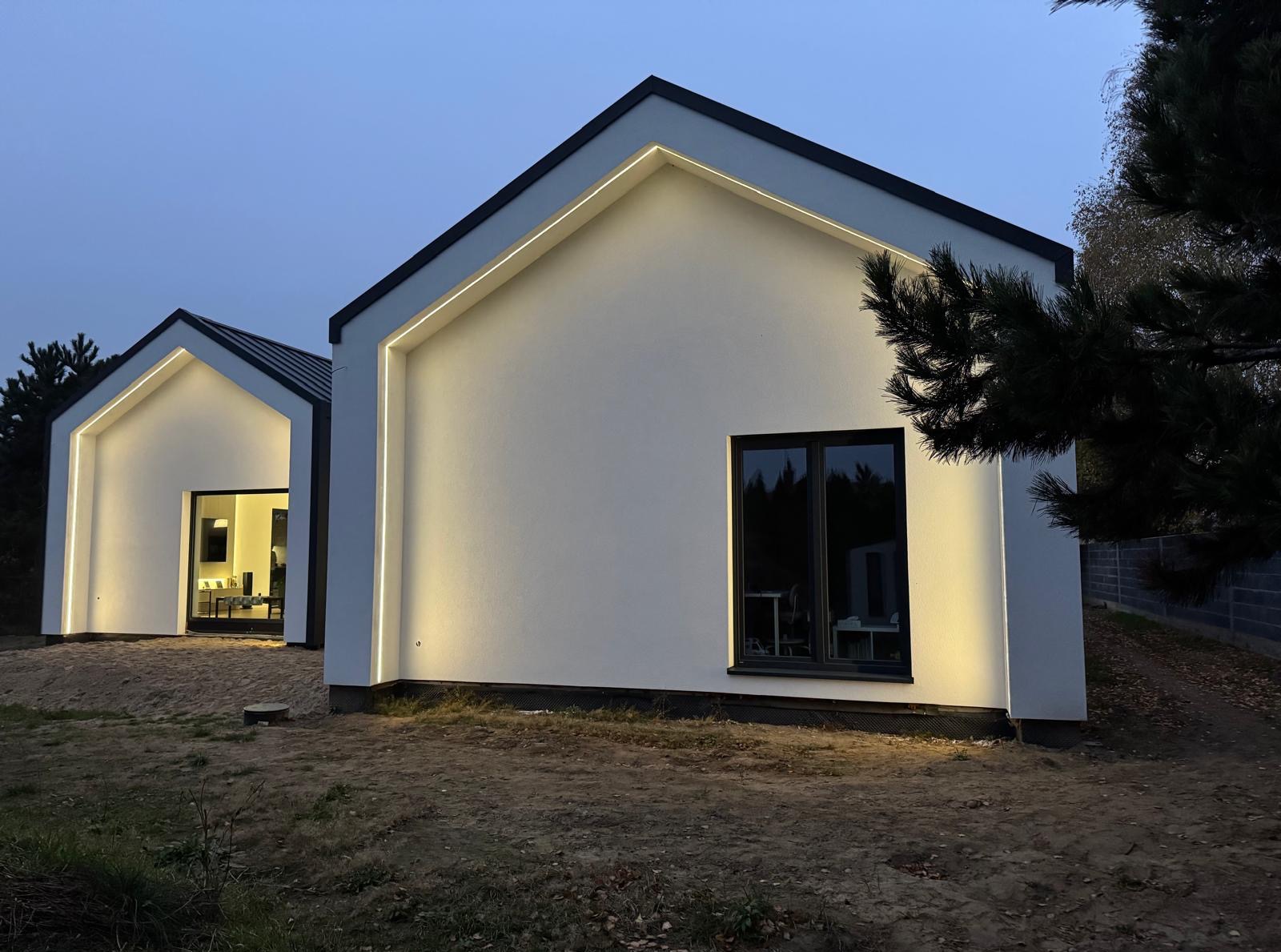 Nowoczesny dom z oświetloną elewacją, widok wieczorny. Białe ściany, czarne okna i dach. Oświetlenie LED podkreśla kształt budynku. Minimalistyczny styl.