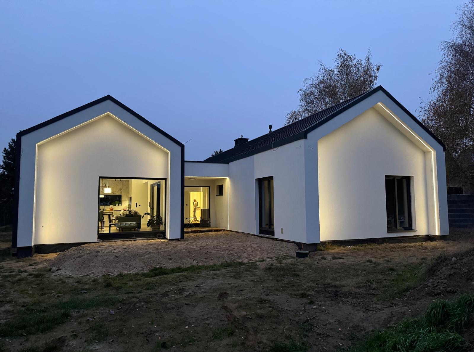 Nowoczesny, parterowy dom z białymi elewacjami i czarnym dachem, oświetlony subtelnym, ledowym światłem, widoczny o zmierzchu.
