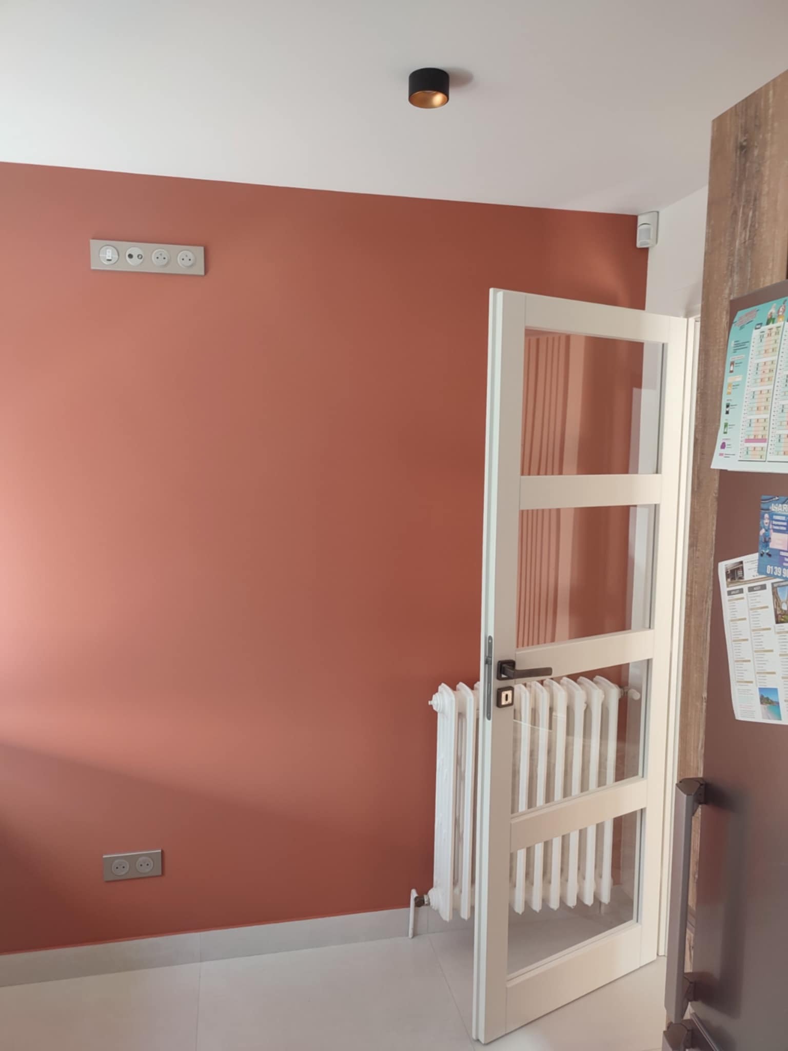 Ściana w odcieniach terakoty z białymi listwami przypodłogowymi, gniazdkami i włącznikami. Białe drzwi z szybami i grzejnik w tle. Minimalistyczne oświetlenie sufitowe.