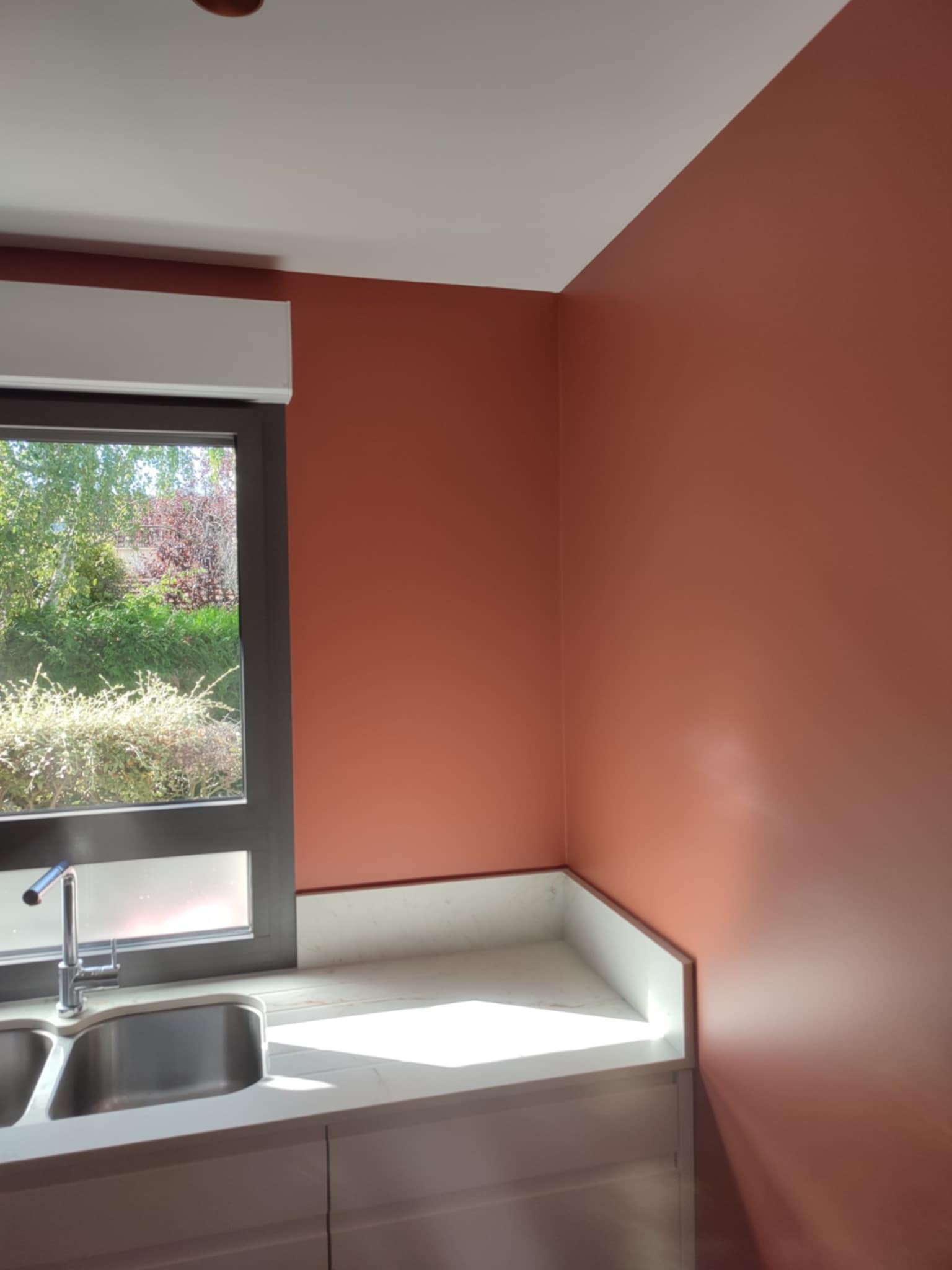 Ściana pomalowana na terakotowy kolor w kuchni z oknem i zlewem. Widok na zieleń za oknem. Gładka, matowa powierzchnia ściany.