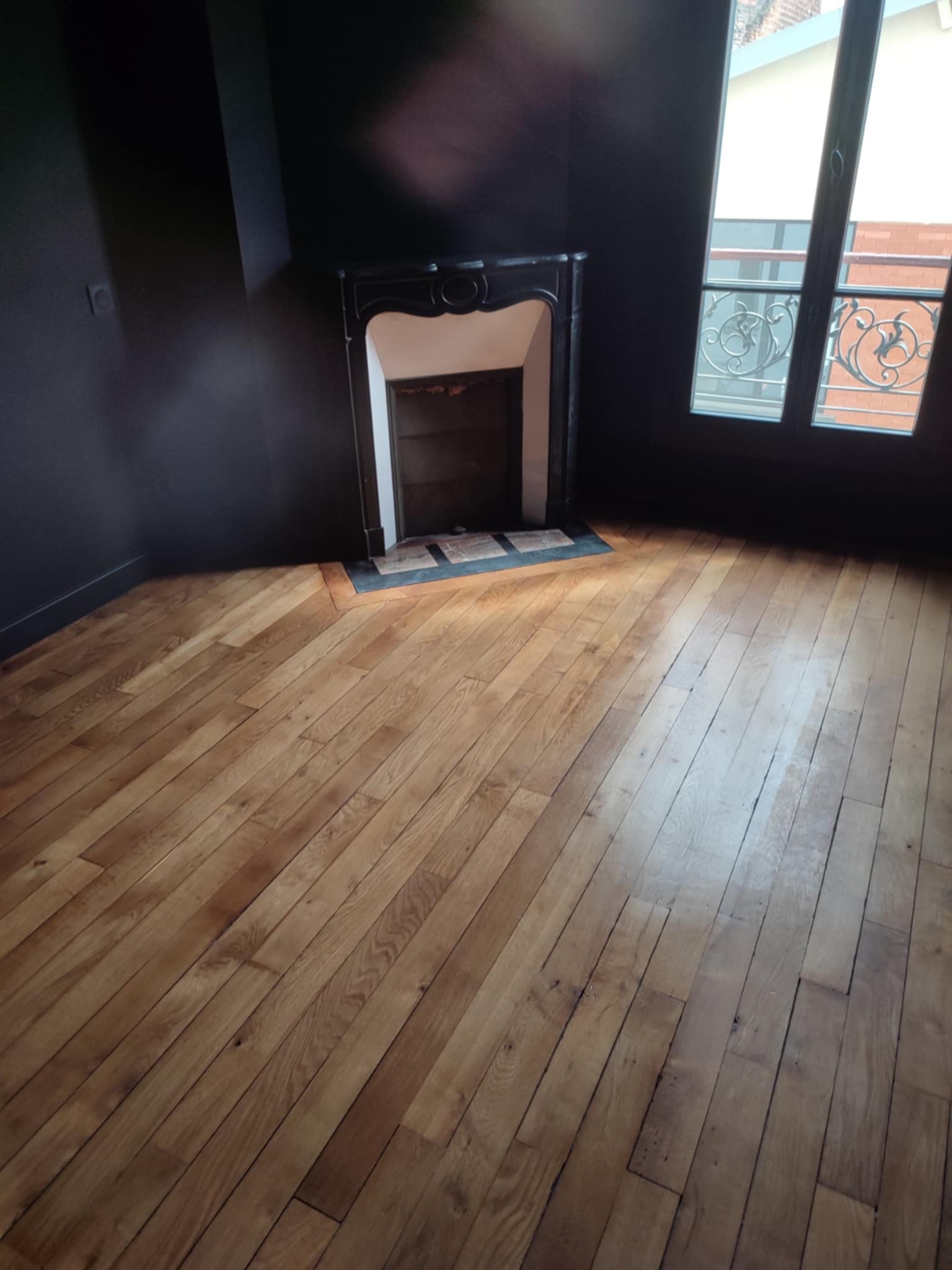 Ciemne ściany i odnowiona drewniana podłoga w pustym pokoju z ozdobnym kominkiem i oknem z ozdobną balustradą. Widok na wnętrze po wykończeniu.