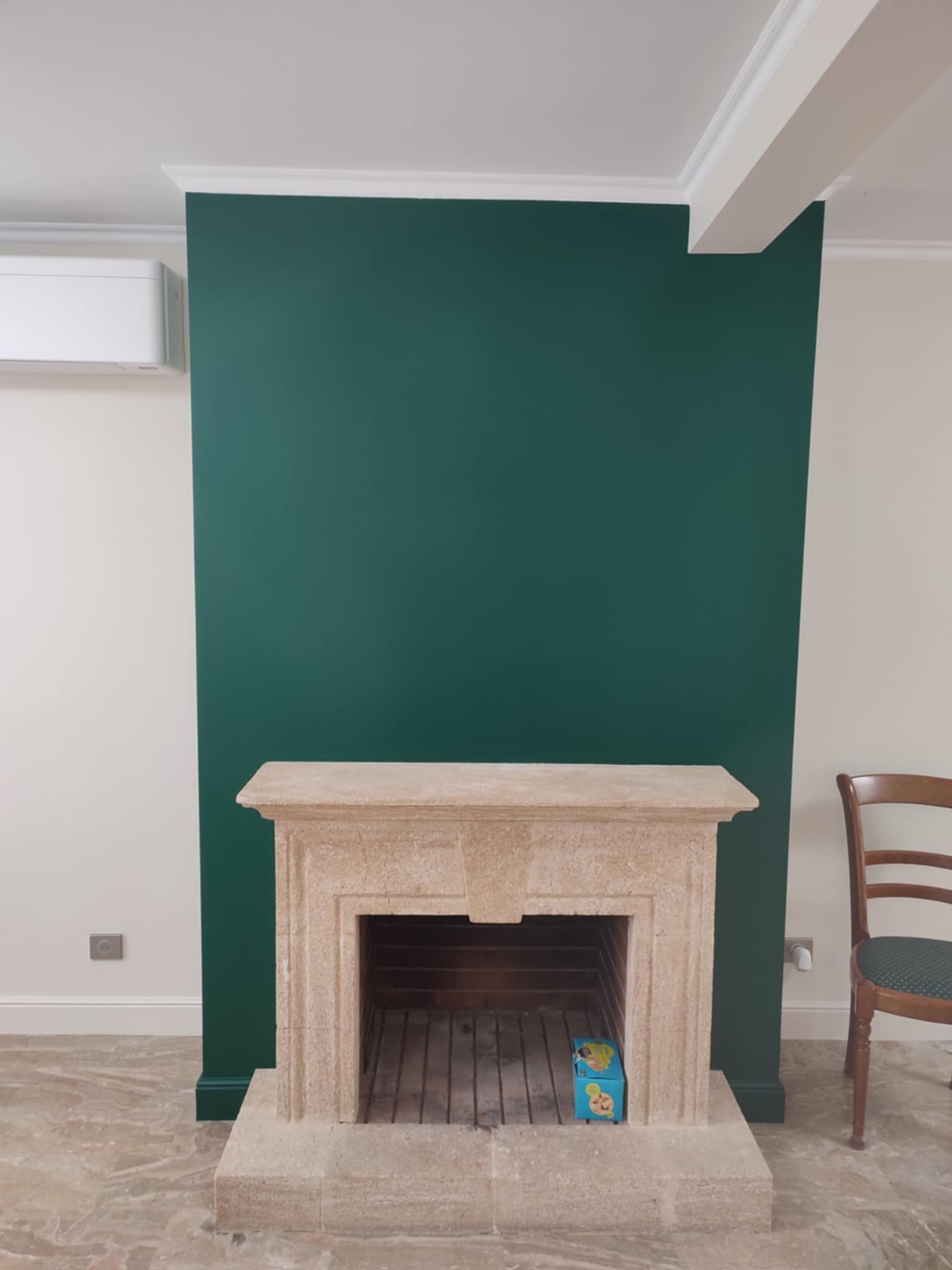 Ściana pomalowana na zielono z kamiennym kominkiem. Widoczny fragment krzesła i klimatyzator. Podłoga marmurowa.