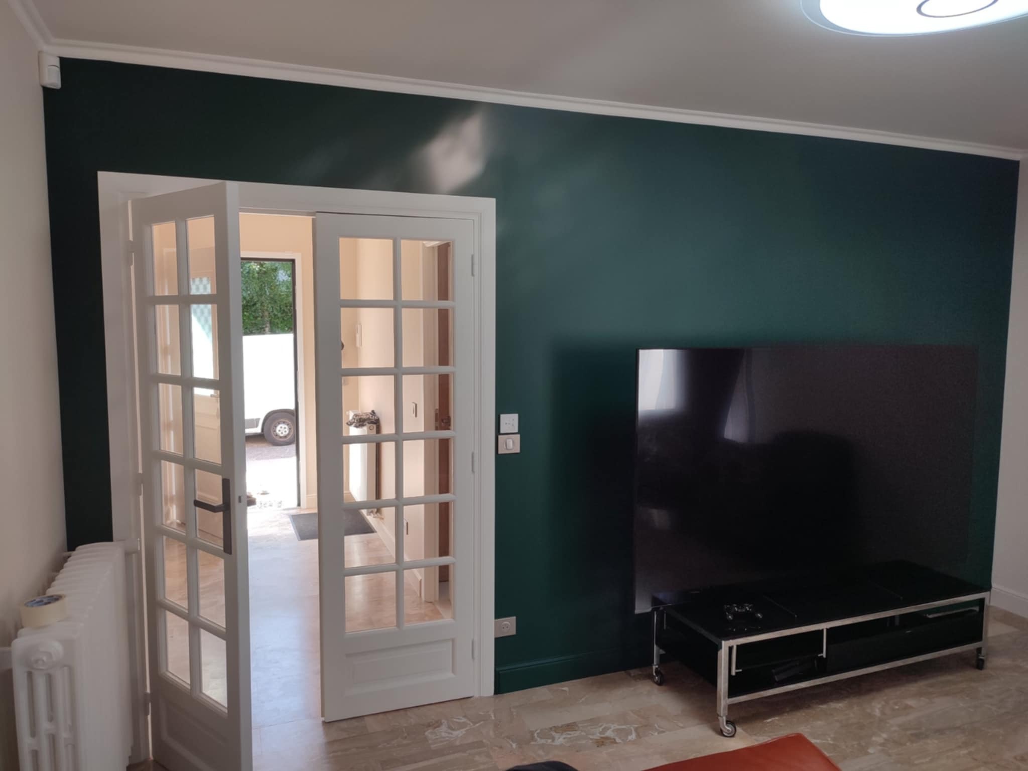 Salon po odświeżeniu: ściana w głębokim odcieniu zieleni, białe, przeszklone drzwi balkonowe, grzejnik z taśmą malarską i nowoczesny stolik RTV.