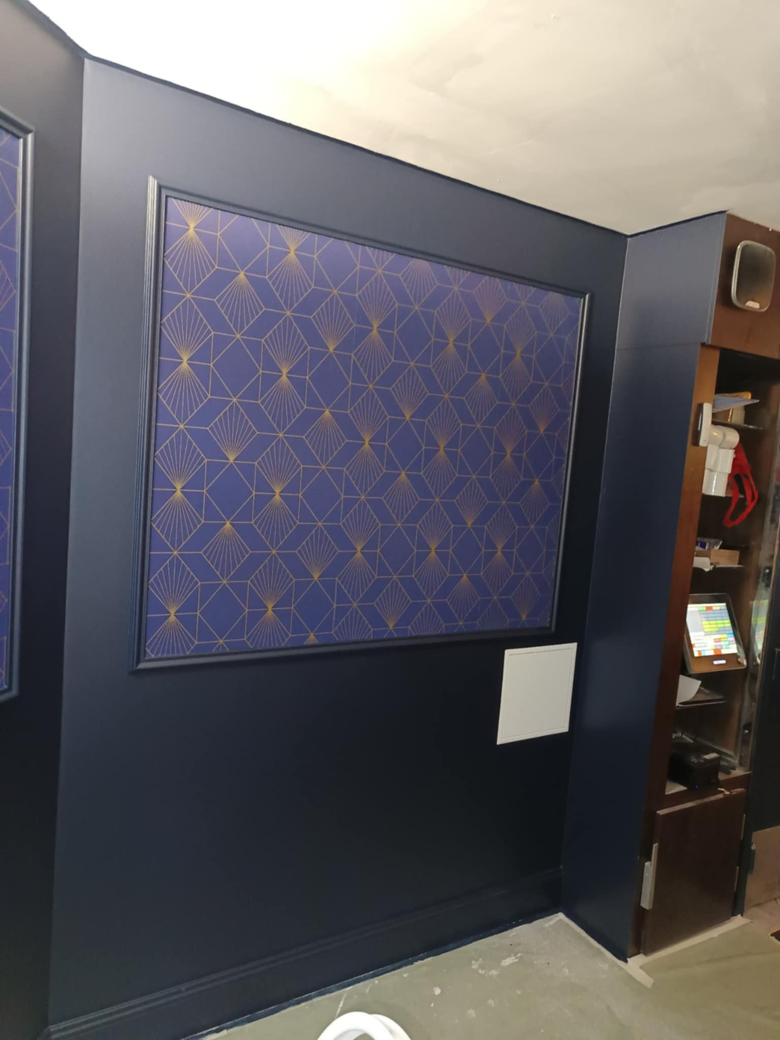 Granatowa ściana z dekoracyjnym panelem w geometryczne wzory, obramowanym czarną listwą. Widoczny fragment szafki i podłogi. Minimalistyczny styl.