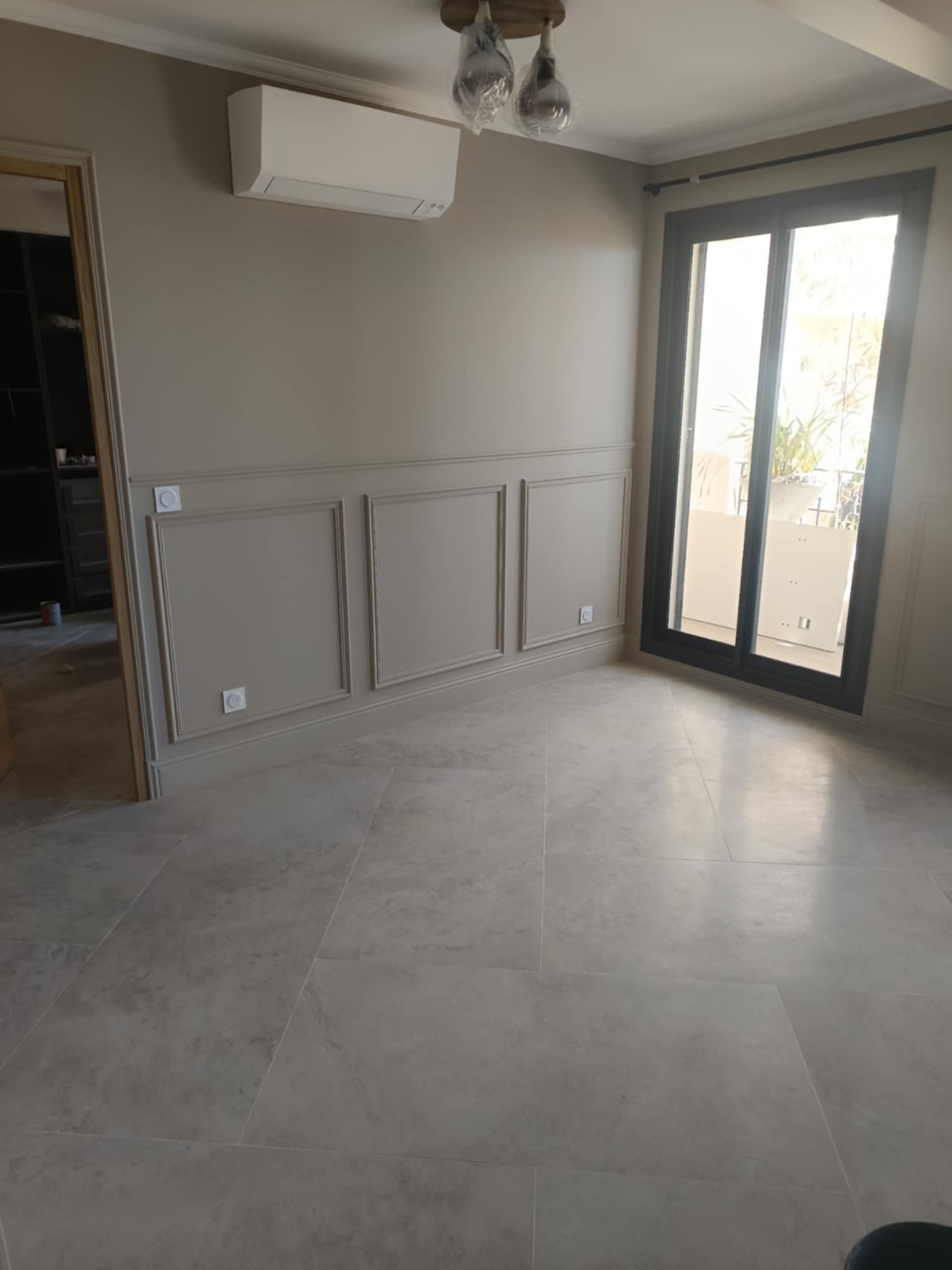 Jasne wnętrze po remoncie: beżowe ściany z dekoracyjnymi panelami, klimatyzator, szare płytki podłogowe, okno balkonowe. Minimalistyczny design.