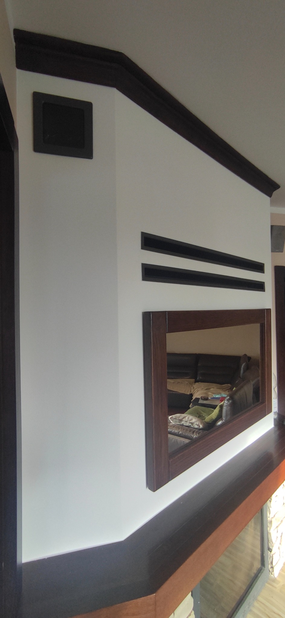 Biała ściana z ciemnobrązowymi elementami dekoracyjnymi: listwa, kratki wentylacyjne i rama lustra. W lustrze odbicie wnętrza pokoju.