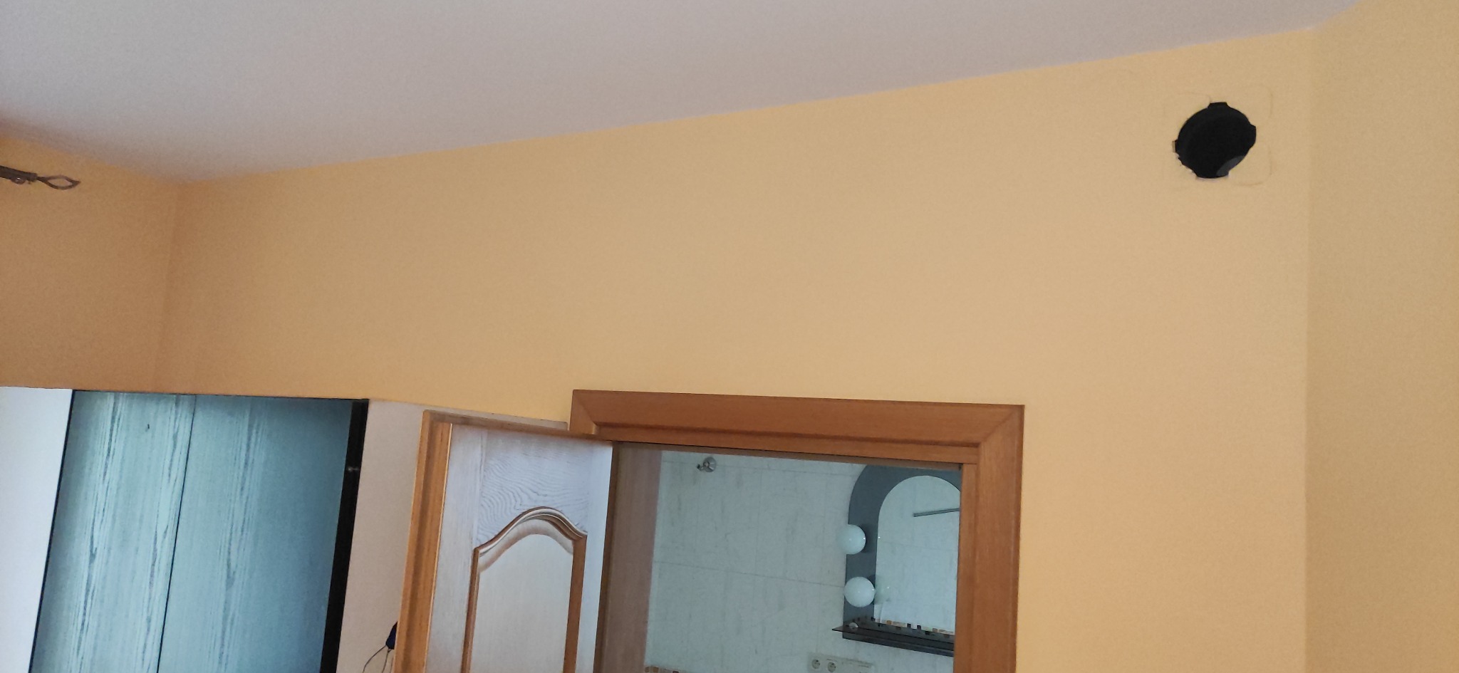 Ściana w odcieniach beżu z widocznym otworem wentylacyjnym i fragmentami ościeżnic drzwiowych. Widoczne ślady po szpachlowaniu wokół otworu.