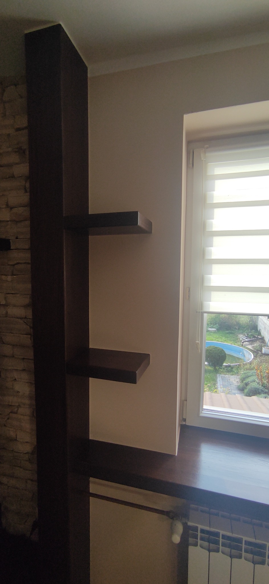 Wykończona wnęka okienna z drewnianym blatem i półkami, obok ściana z dekoracyjną okładziną kamienną. Widok na ogród przez okno z roletami.