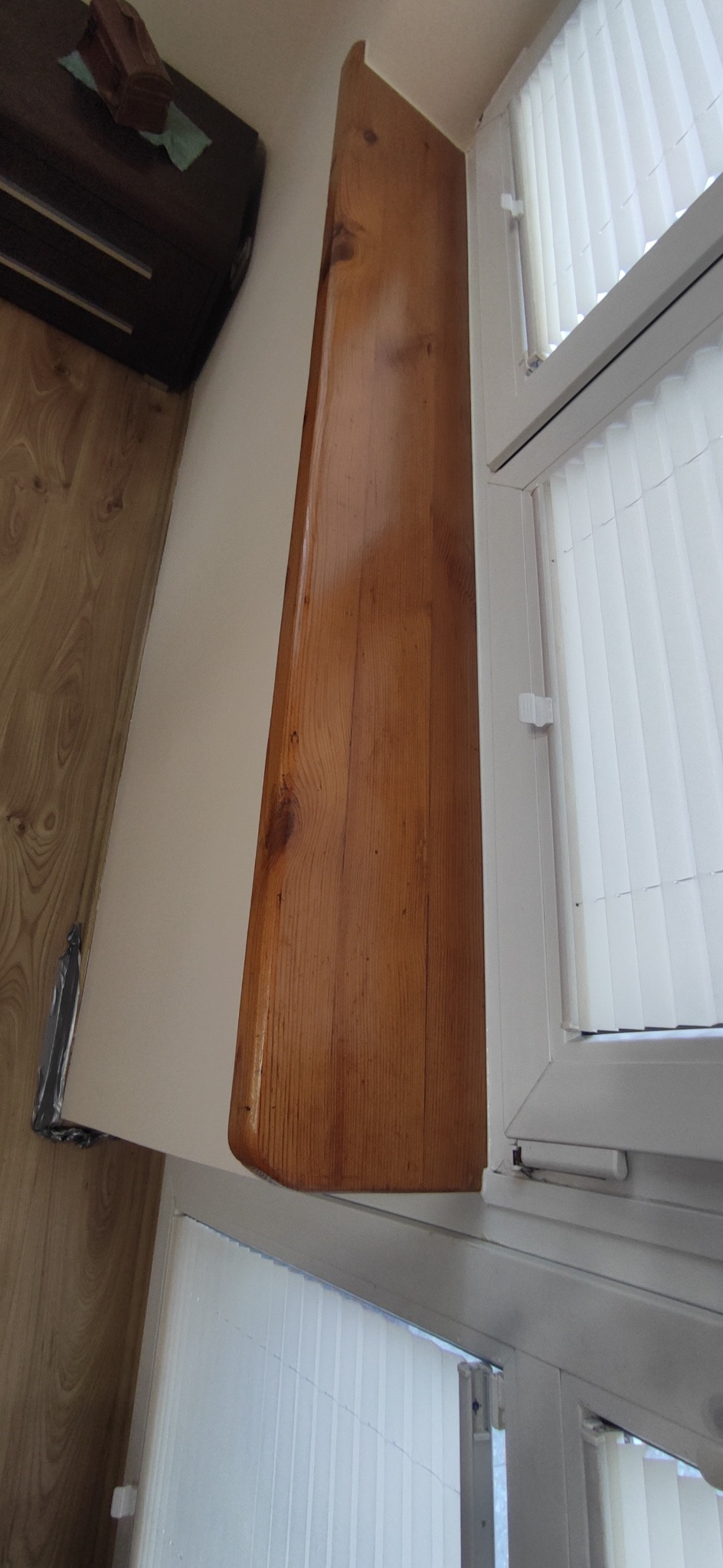 Drewniana listwa wykończeniowa przy oknie z żaluzjami, zamontowana na pomalowanej ścianie. Widoczne słoje drewna i fragment podłogi.