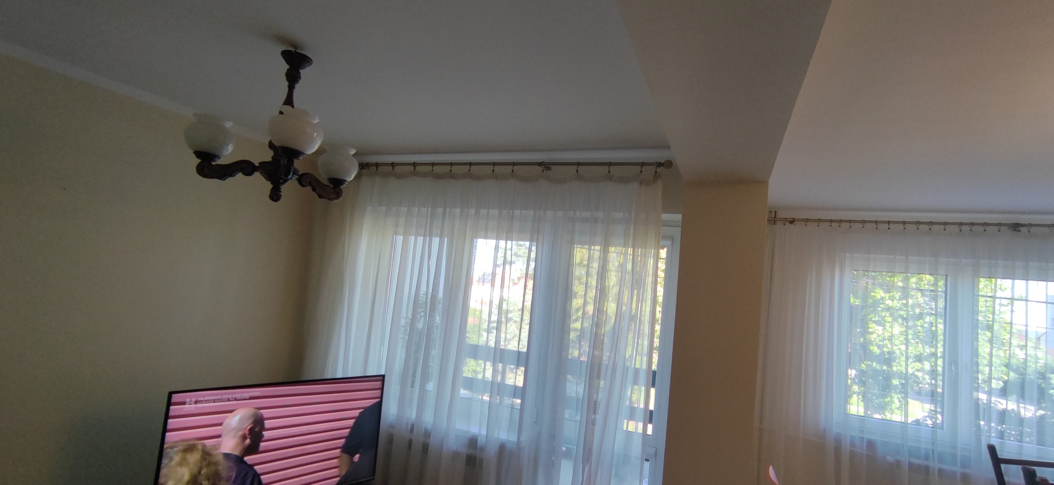 Jasne wnętrze pokoju z kremowymi ścianami, białymi firankami w oknach i klasycznym żyrandolem. Widoczny fragment telewizora. Ujęcie z dołu.