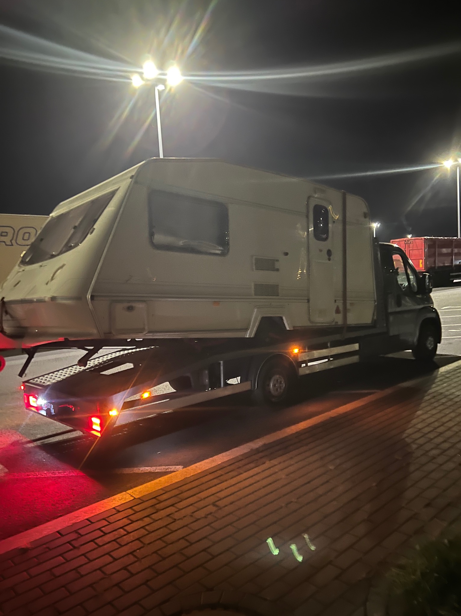 Nocny transport przyczepy kempingowej na niskopodwoziowej platformie, oświetlonej światłami ulicznymi i czerwonymi światłami lawety. Widok z boku, ciemne tło.