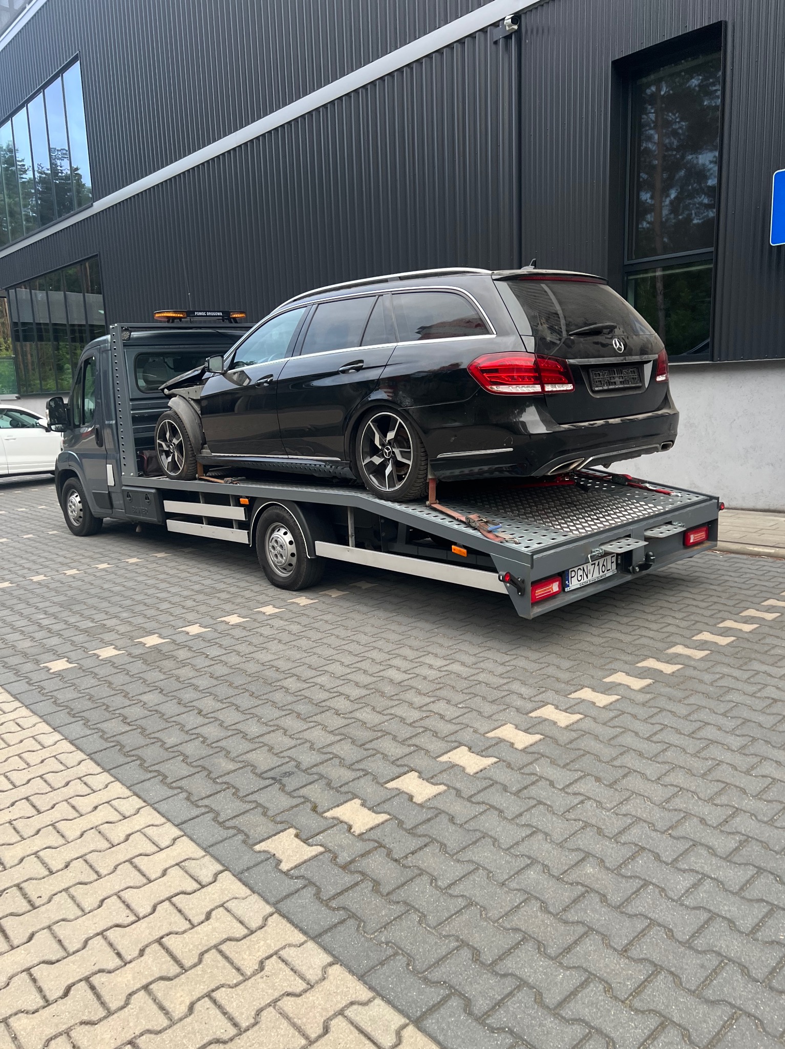 Czarny Mercedes kombi z uszkodzonym przodem na szarej lawecie pomocy drogowej, zaparkowanej na kostce brukowej przed nowoczesnym, ciemnym budynkiem.