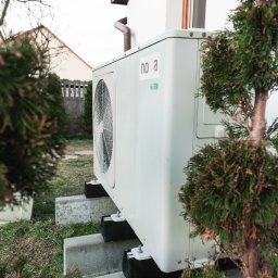 ENERGIA PLUS GAZ SPÓŁKA Z OGRANICZONĄ ODPOWIEDZIALNOŚCIĄ - Boczny widok pompy ciepła Nova, zamontowanej na betonowych blokach przy białym domu z brązowymi detalami, otoczonej zielenią. Widoczny wentylator i naklejka z oznaczeniem.
