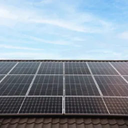 Panele słoneczne na brązowym dachu pod błękitnym niebem. Widok z dołu, podkreślający regularny układ paneli i ich integrację z architekturą budynku.