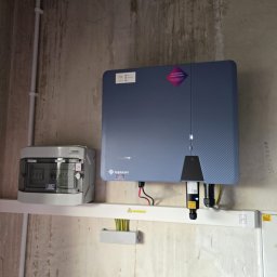 ENERGIA PLUS GAZ SPÓŁKA Z OGRANICZONĄ ODPOWIEDZIALNOŚCIĄ - Instalacja fotowoltaiczna Solplanet: inwerter na ścianie z widoczną skrzynką rozdzielczą i okablowaniem. Ściana betonowa, widoczne ślady wilgoci.