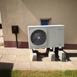 ENERGIA PLUS GAZ SPÓŁKA Z OGRANICZONĄ ODPOWIEDZIALNOŚCIĄ - Biała pompa ciepła Nova, zamontowana przy beżowej ścianie budynku z brązowym cokołem, na betonowych wspornikach. Widoczna skrzynka gazowa.
