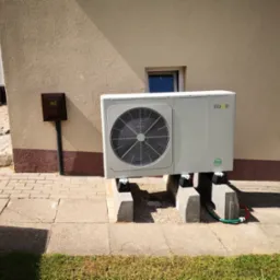 Biała pompa ciepła Nova, zamontowana przy beżowej ścianie budynku z brązowym cokołem, na betonowych wspornikach. Widoczna skrzynka gazowa.