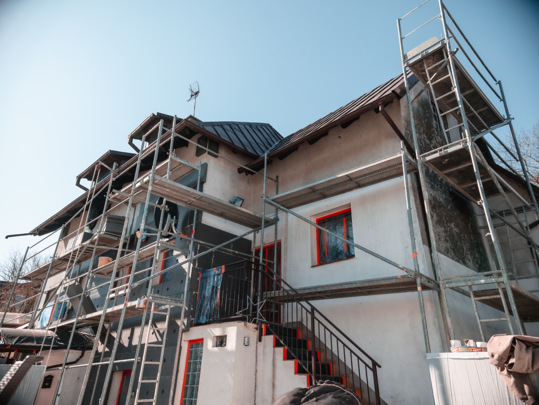 Ocieplanie budynku styropianem na rusztowaniu w Sopocie. Widoczne okna zabezpieczone folią i częściowo ocieplona elewacja. Prace w toku, widok z dołu.