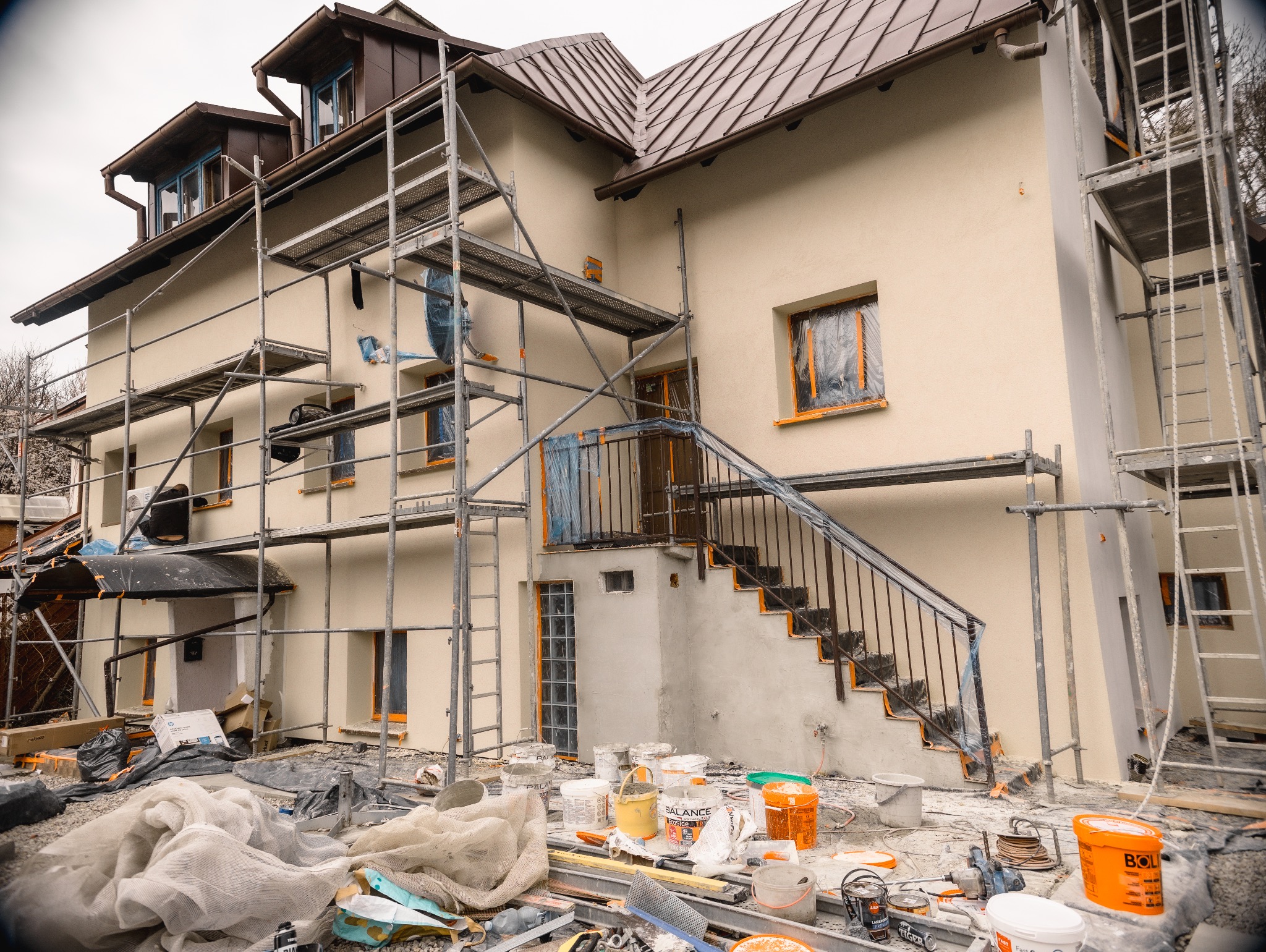 Dom w trakcie elewacji: rusztowania, zabezpieczone okna folią, farby, narzędzia. Budynek z brązowym dachem i beżową fasadą w trakcie prac wykończeniowych.