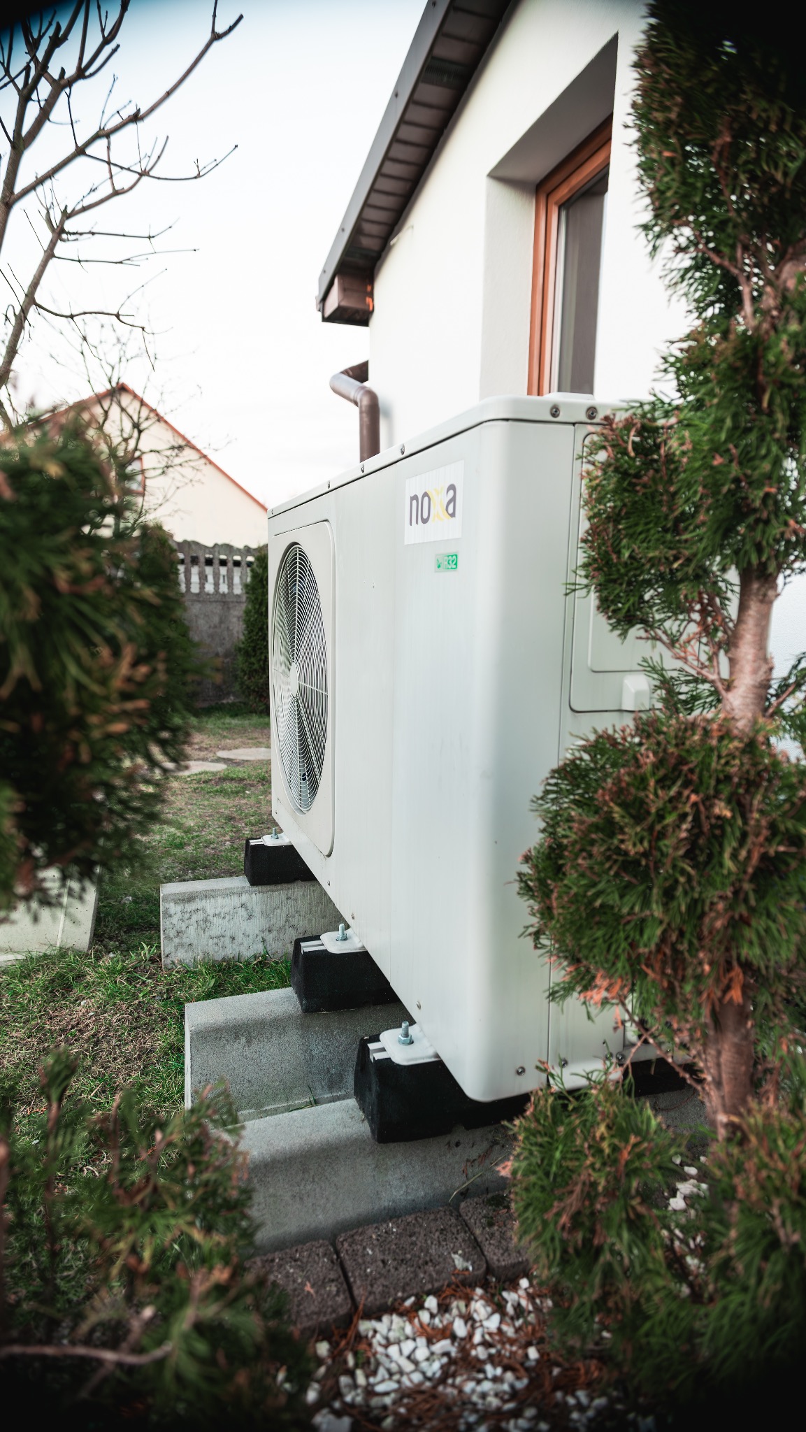 Boczny widok pompy ciepła Nova, zamontowanej na betonowych blokach przy białym domu z brązowymi detalami, otoczonej zielenią. Widoczny wentylator i naklejka z oznaczeniem.