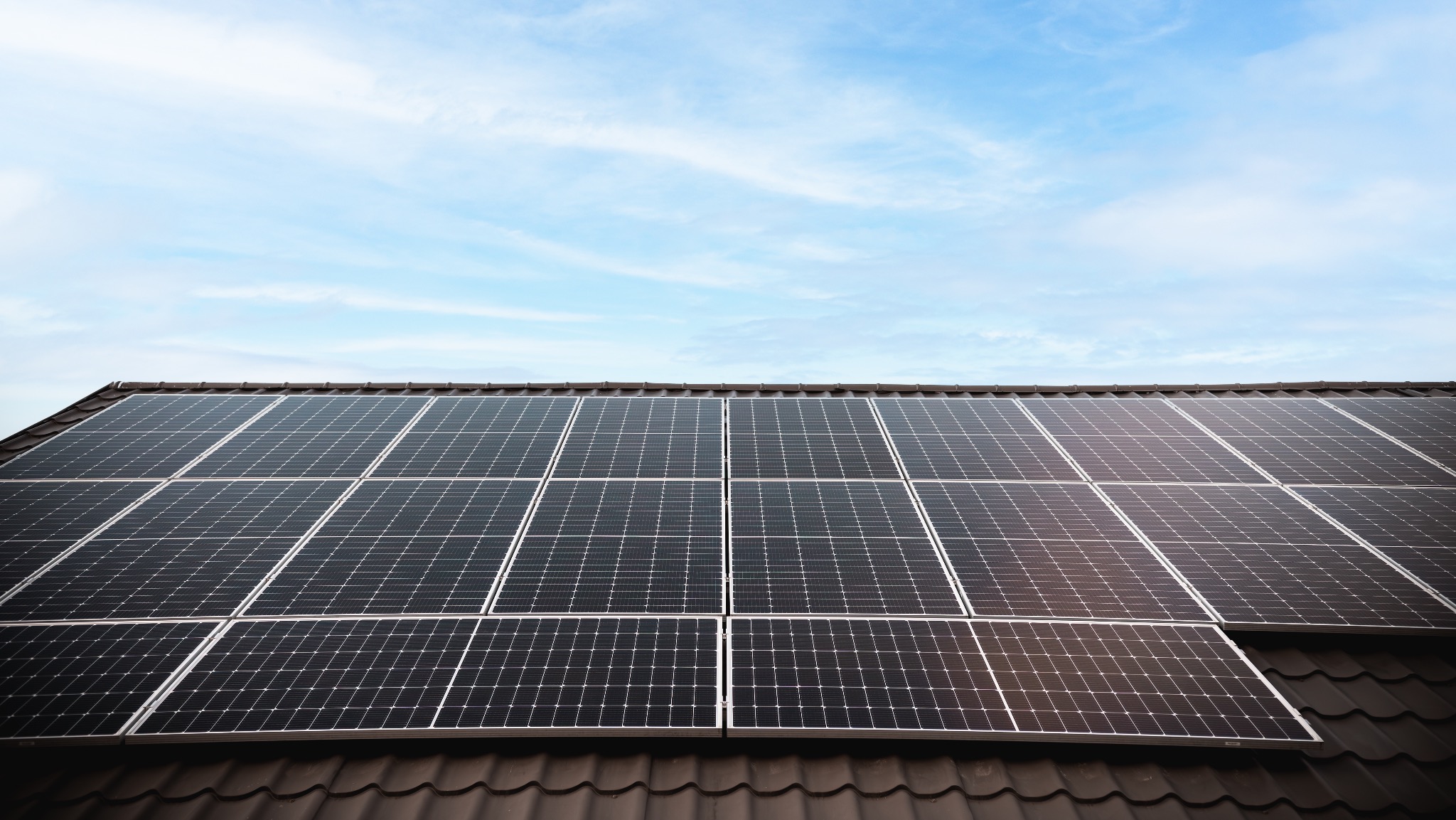 Panele słoneczne na brązowym dachu pod błękitnym niebem. Widok z dołu, podkreślający regularny układ paneli i ich integrację z architekturą budynku.