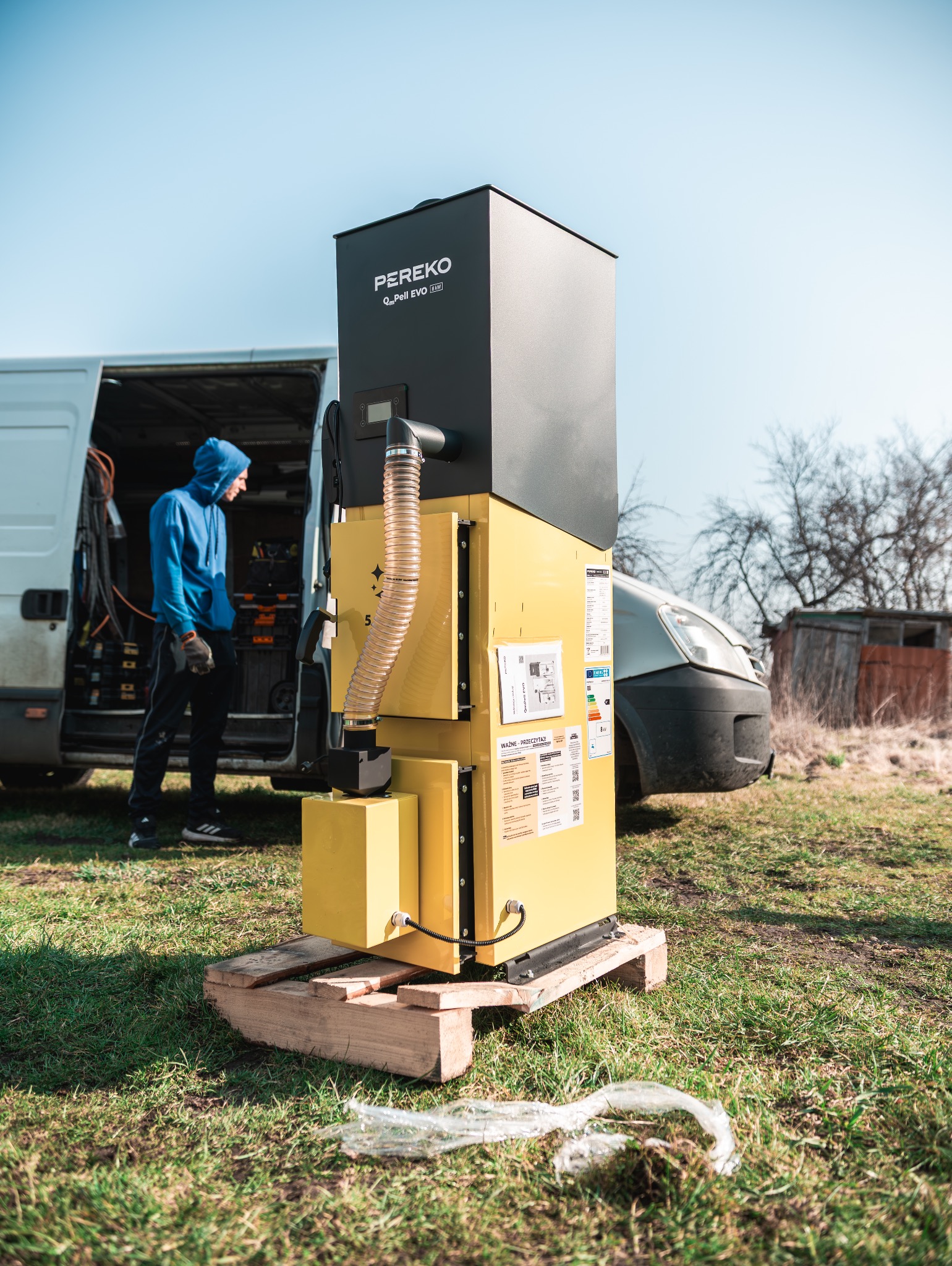 Żółto-czarny piec na pellet PEREKO QmPell EVO 30 kW na palecie, z otwartym busem w tle, w trakcie montażu lub dostawy. Widoczny pracownik w niebieskiej bluzie.
