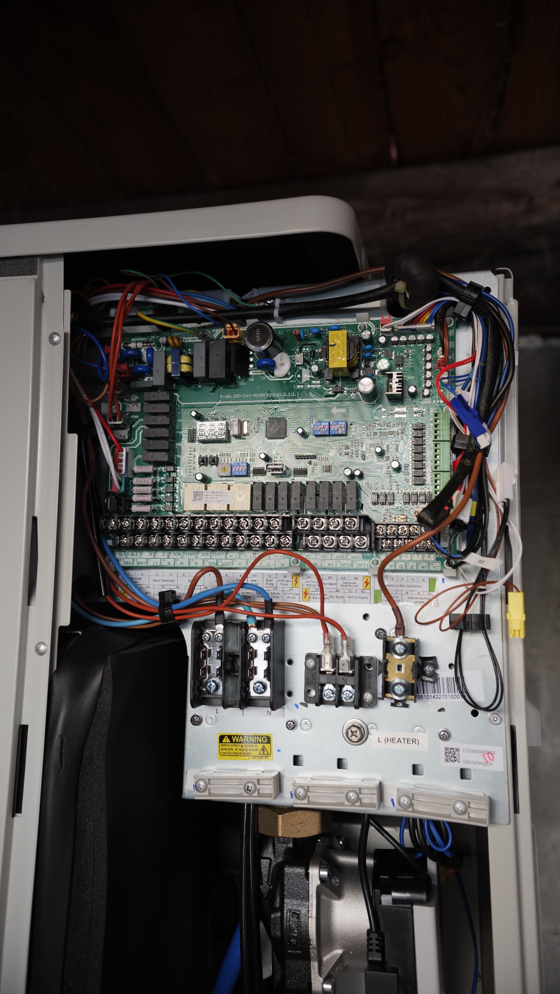 Otwarta obudowa pompy ciepła z widoczną płytą sterującą, licznymi przewodami i elementami elektronicznymi. Widoczne złącza, kable w różnych kolorach oraz ostrzeżenie.