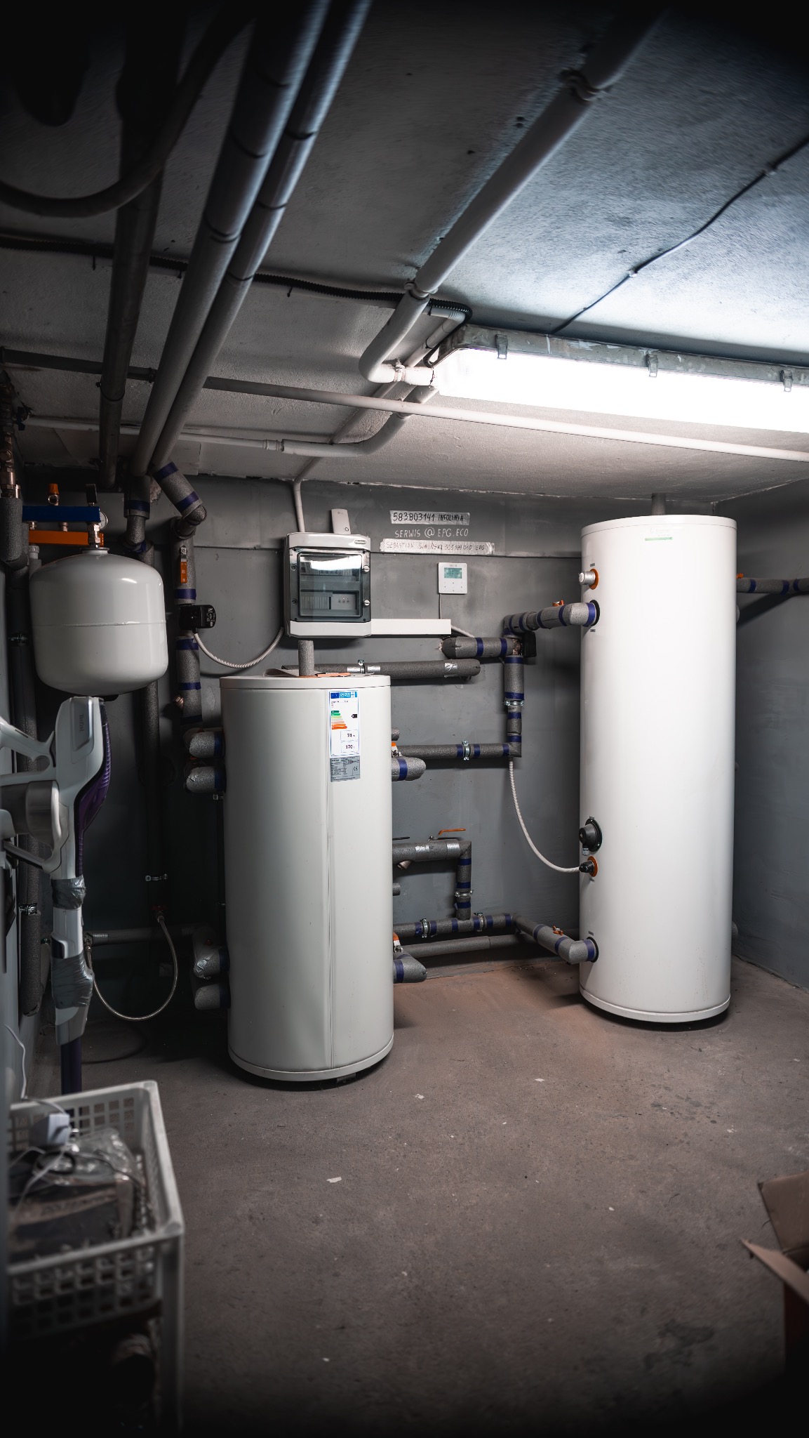 Dwa białe zbiorniki akumulacyjne w piwnicy z szarymi ścianami i rurami. Instalacja z izolowanymi rurami. Widoczna skrzynka elektryczna na ścianie.