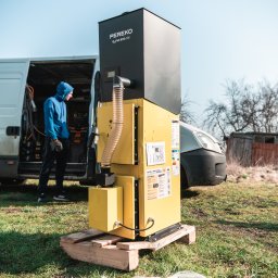 IQDOM 24 - Żółto-czarny piec na pellet PEREKO QuPell EVO RAW na palecie, obok bus instalatora. Widoczne etykiety informacyjne i przewód. Montaż w terenie zielonym.