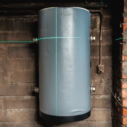 IQDOM 24 - Szary akumulator ciepła zamontowany na ceglanej ścianie, wyznaczony laserowo. Widoczne rury i kable w surowym pomieszczeniu.