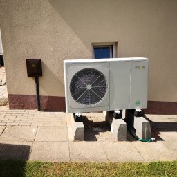 IQDOM 24 - Biała pompa ciepła Nova zamontowana na betonowych blokach przy budynku z brązową skrzynką gazową. Widoczny fragment trawnika i chodnika.