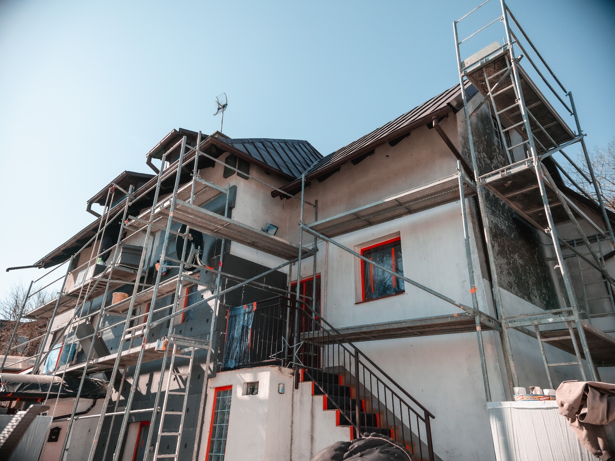 Dom w trakcie ocieplania, z rusztowaniami. Część ściany pokryta izolacją, okna zabezpieczone folią. Widoczne materiały budowlane.