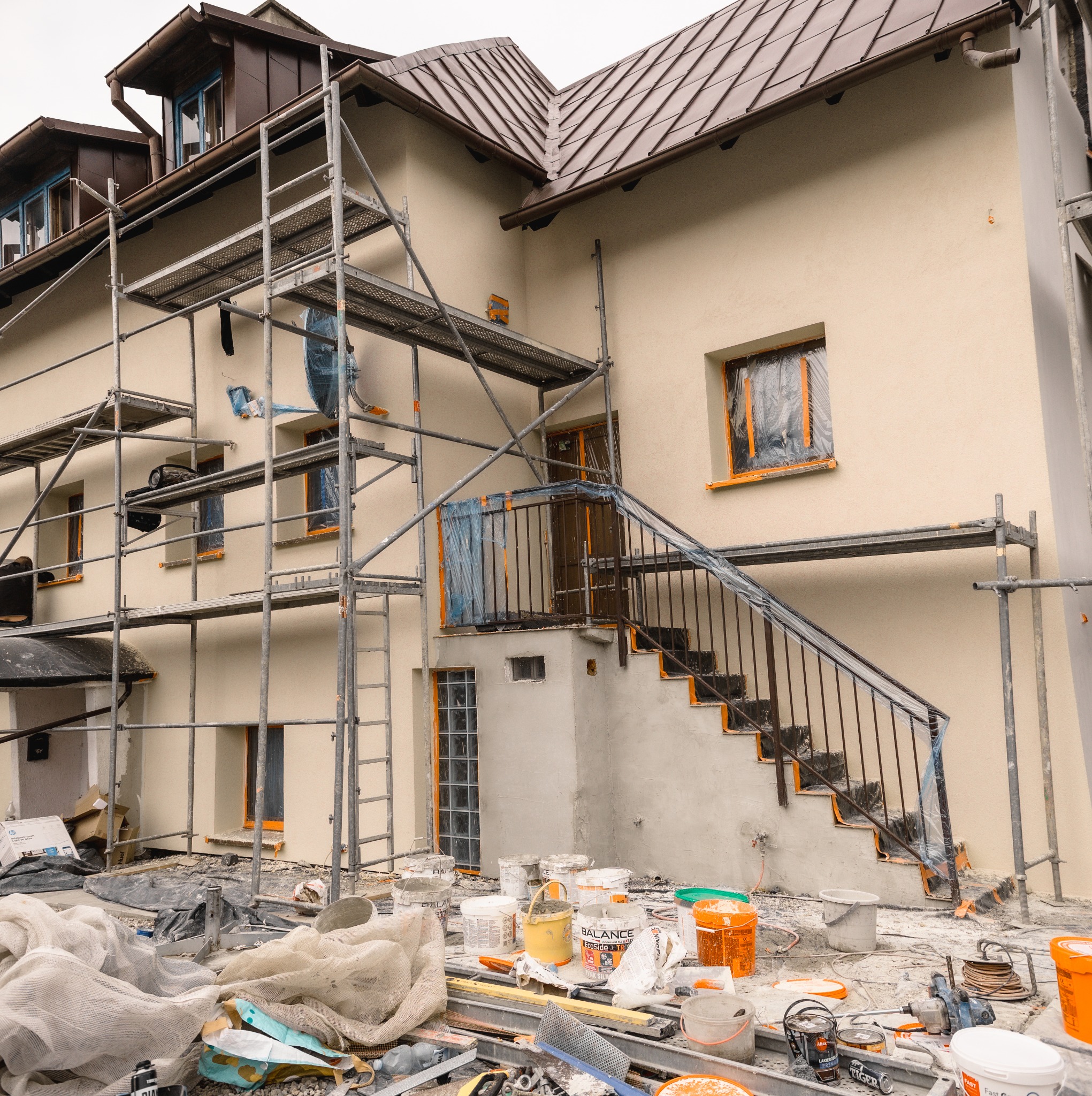 Elewacja domu w trakcie prac wykończeniowych. Widoczne rusztowania, farby, zabezpieczone okna i schody. Jasna elewacja, brązowy dach.