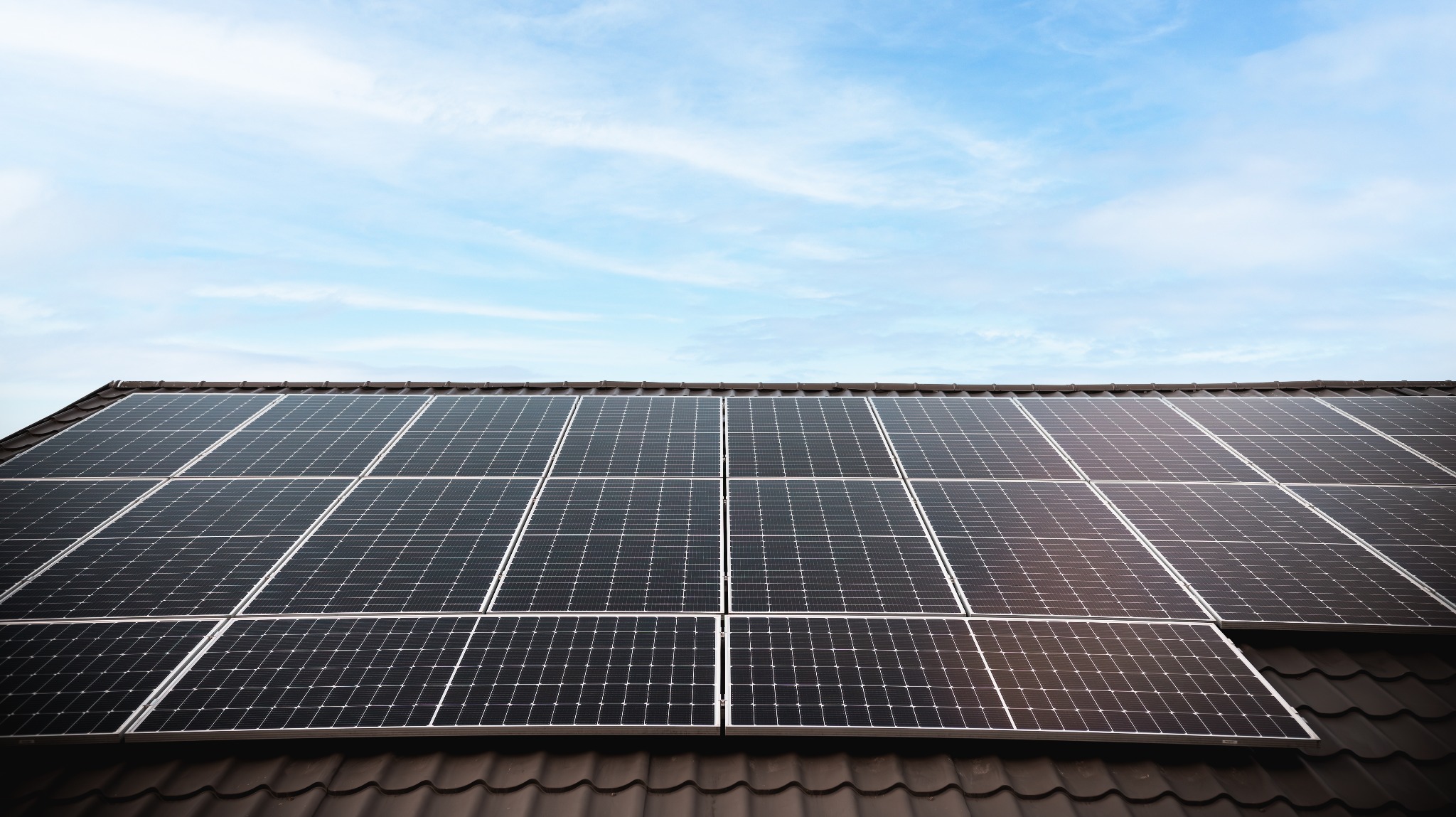 Panele słoneczne na ciemnym, spadzistym dachu z brązowej dachówki pod błękitnym niebem z delikatnymi chmurami. Widok z dołu, lekko z prawej strony.