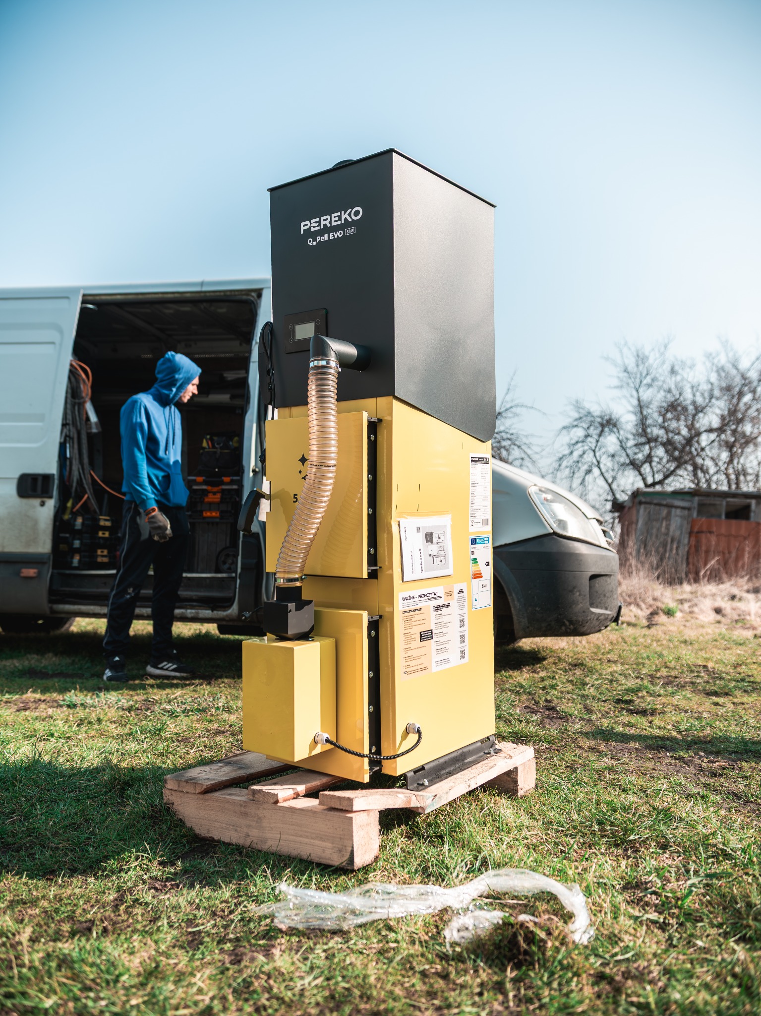 Żółto-czarny piec na pellet PEREKO QuPell EVO RAW na palecie, obok bus instalatora. Widoczne etykiety informacyjne i przewód. Montaż w terenie zielonym.
