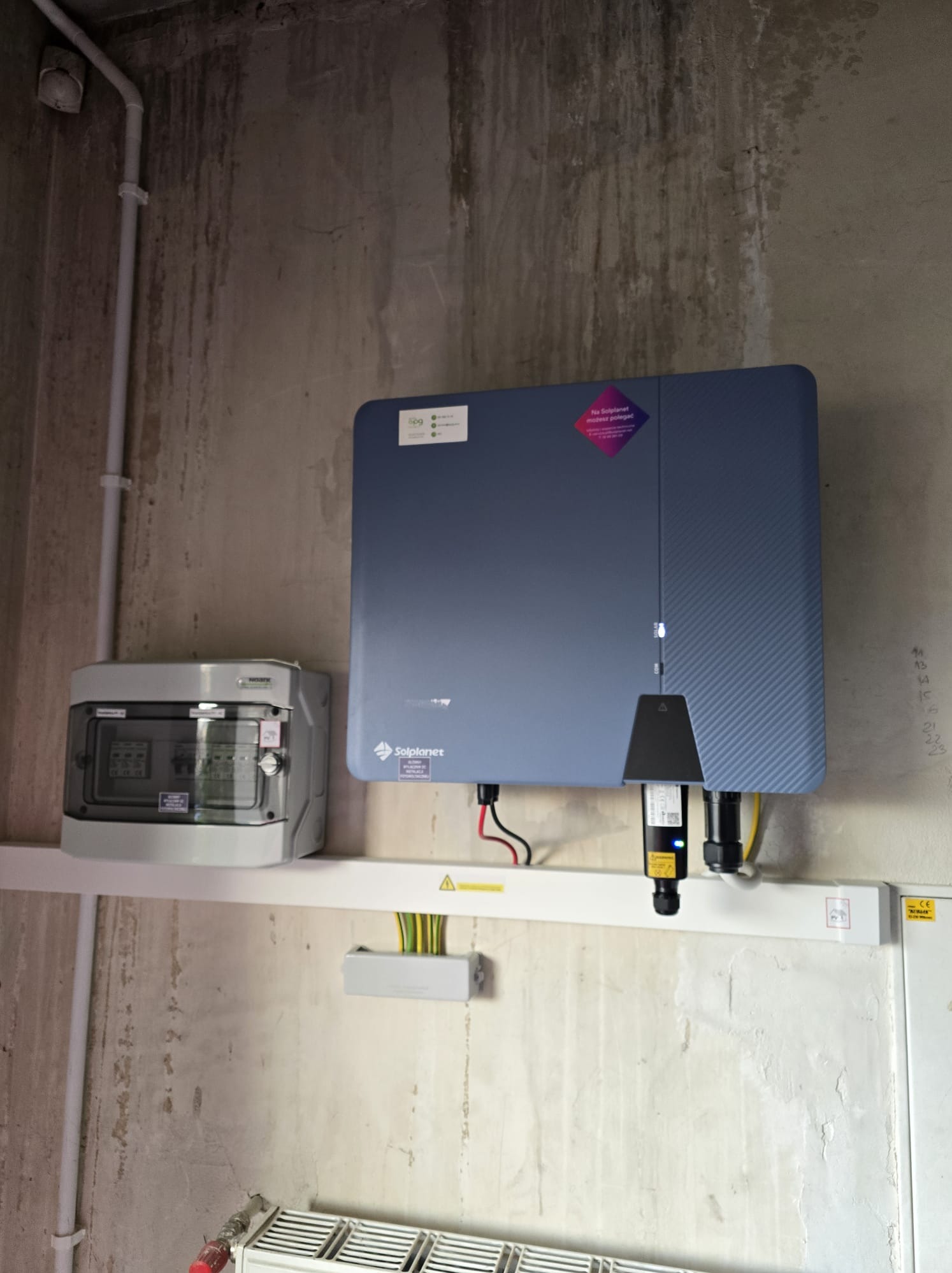 Instalacja falownika Solplanet z rozdzielnicą na ścianie o surowym wykończeniu. Widoczne kable i elementy instalacji elektrycznej.