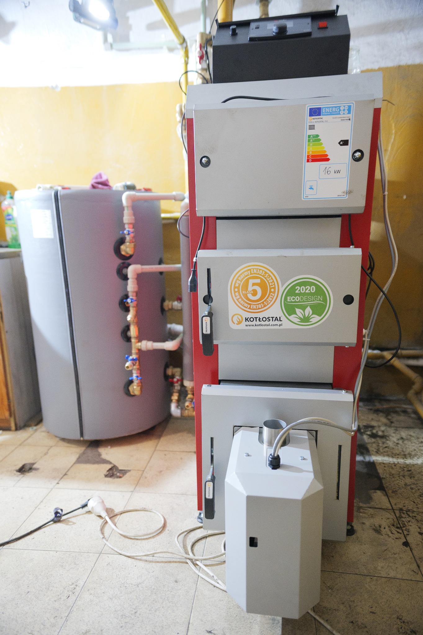 Kocioł na paliwo stałe marki Kotłostal, z etykietą energetyczną i oznaczeniem Ecodesign 2020, obok zbiornika akumulacyjnego, w pomieszczeniu kotłowni.