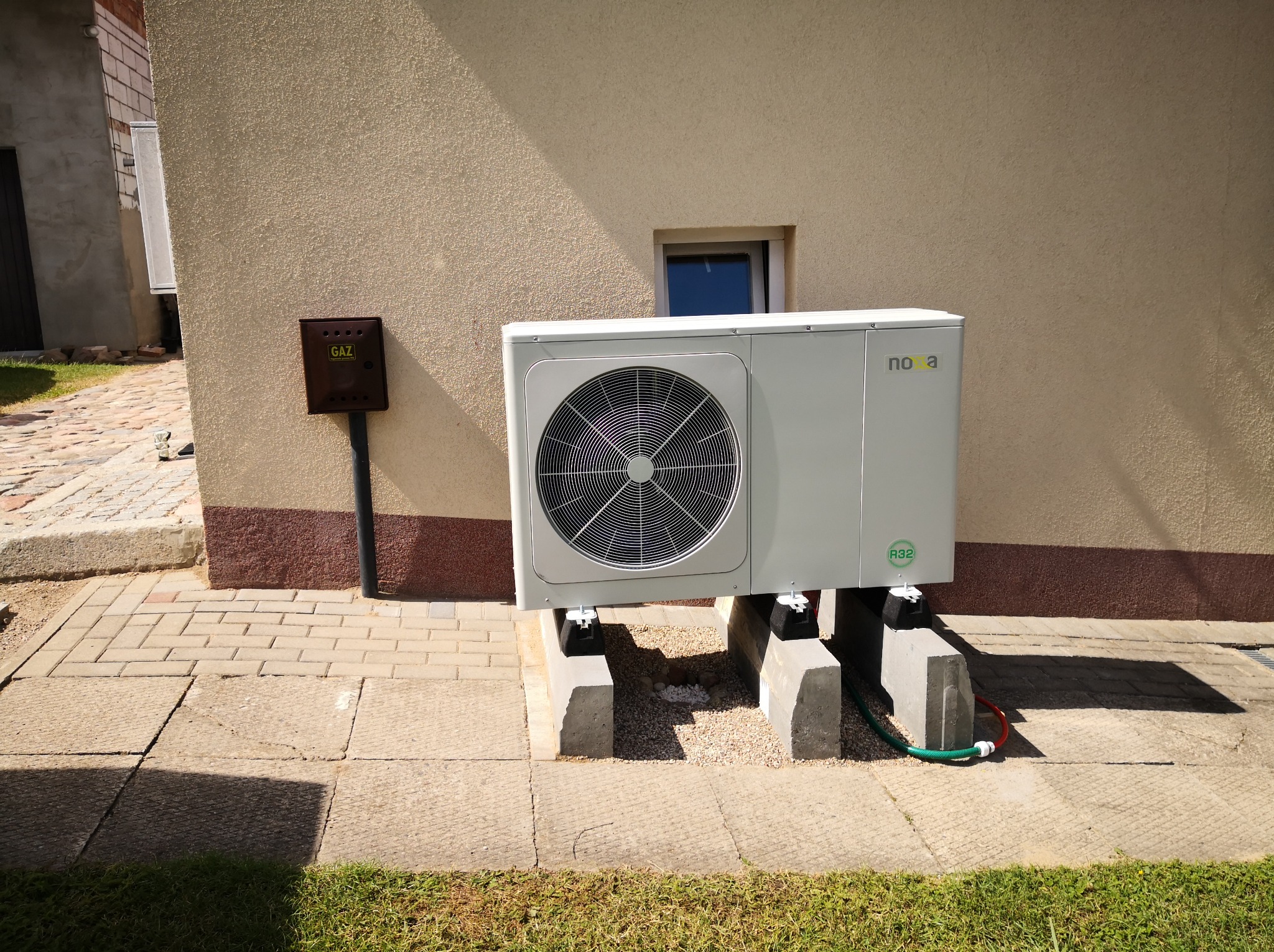 Biała pompa ciepła Nova zamontowana na betonowych blokach przy budynku z brązową skrzynką gazową. Widoczny fragment trawnika i chodnika.
