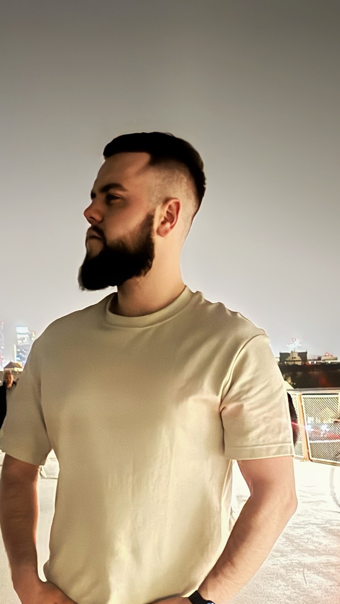 Mężczyzna z brodą i modną fryzurą, patrzy w lewo, na tle miejskiej panoramy nocą. Widoczne detale strzyżenia i pielęgnacji zarostu.